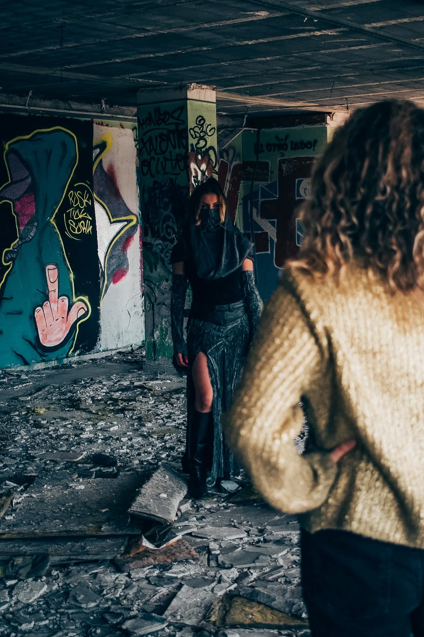 People in a graffiti-covered space with rubble on the ground; one person in the foreground has curly hair and is wearing a tan sweater, while a woman in the background has long hair, sunglasses, a black top, and ripped jeans.