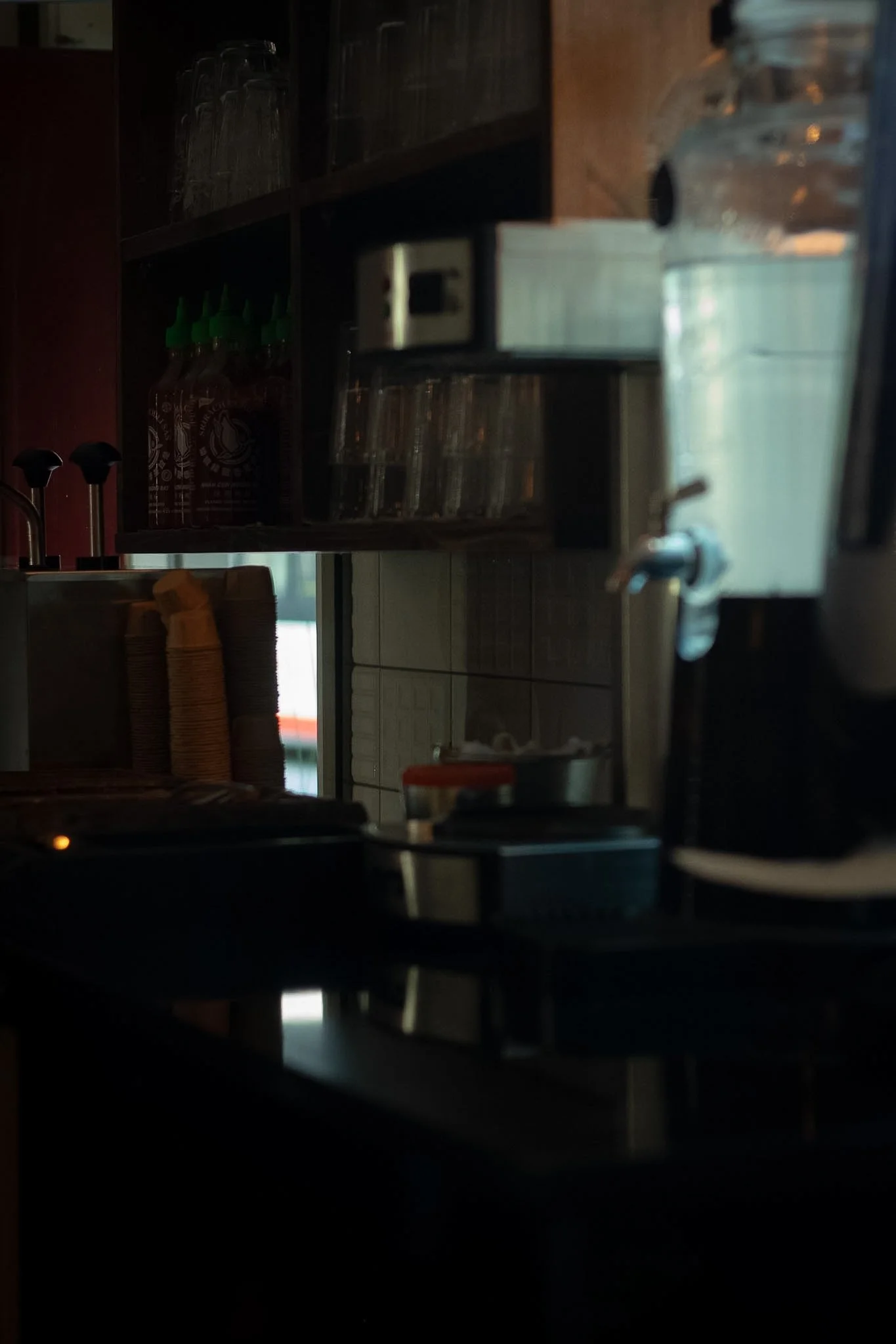 A dimly lit indoor scene of a coffee shop or kitchen area, with shelves holding glasses, a stack of disposable cups, and various kitchen items visible in the background.