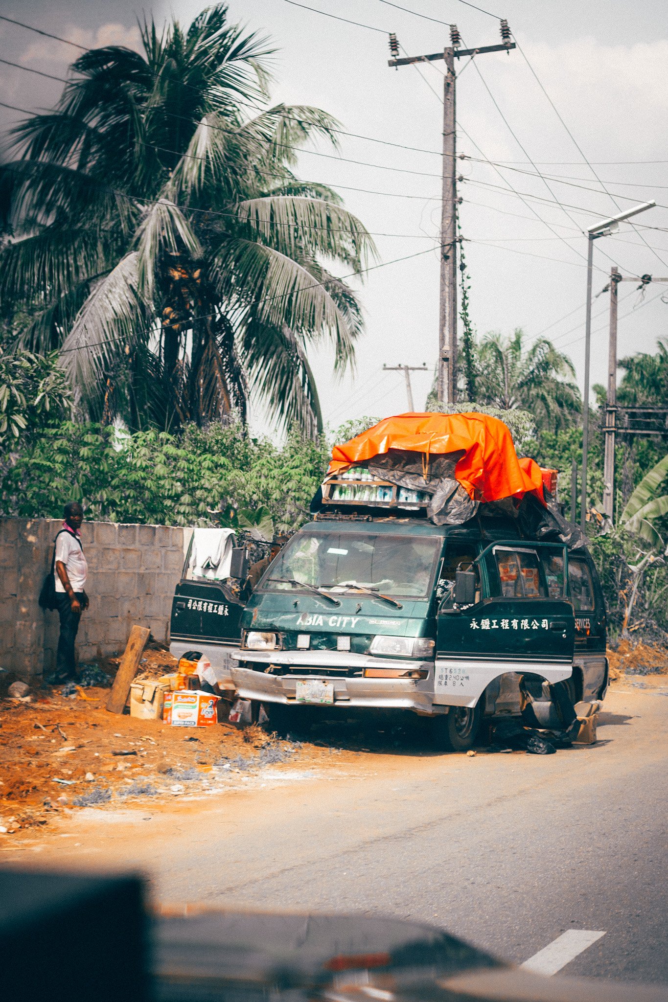 A street scene with a black vehicle labeled 'Asia City' in front of a stone wall, with a man standing nearby. There are trees, utility poles, and wires in the background. The ground is dirt, and there are boxes or packages on the ground near the vehi