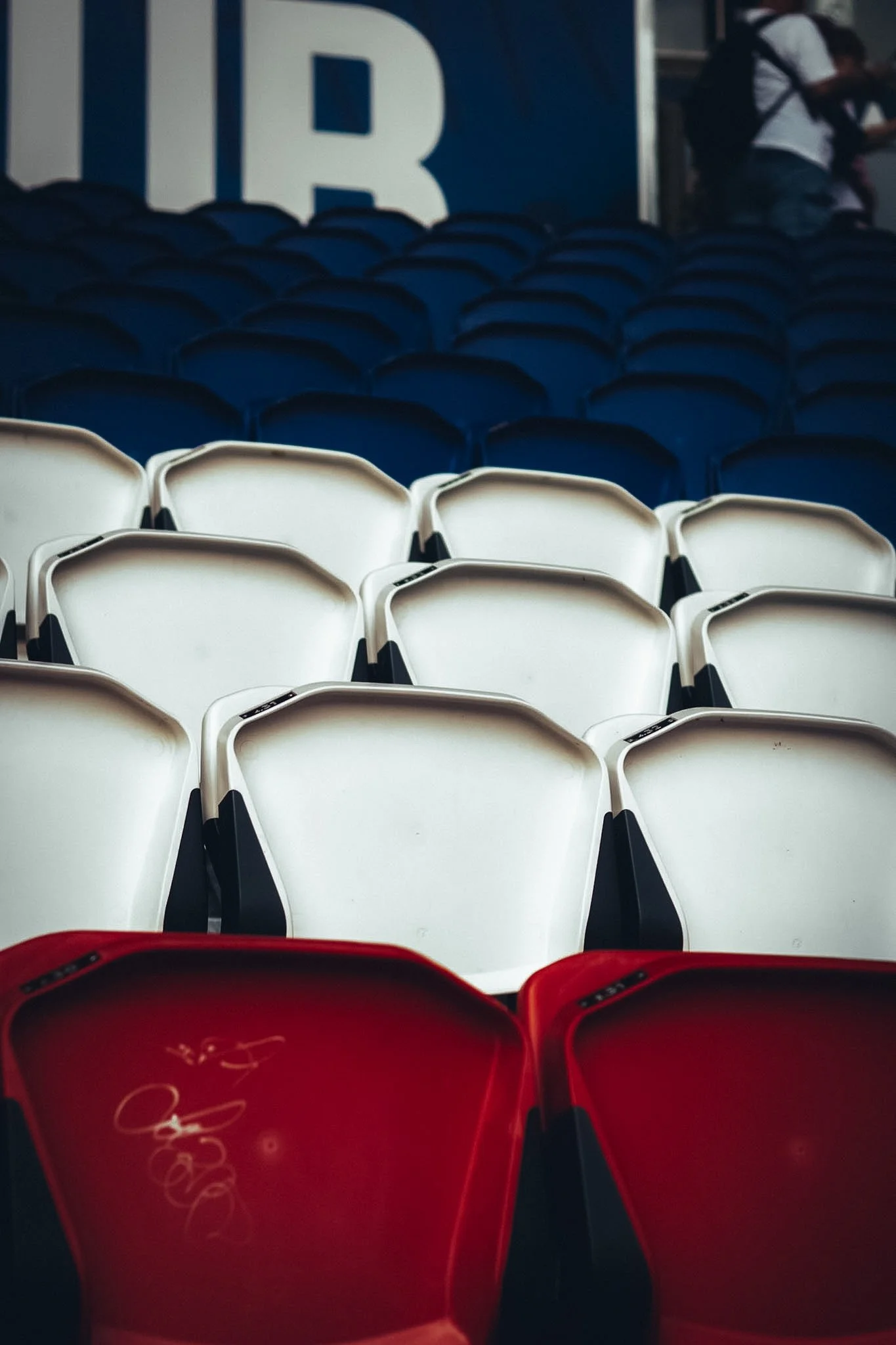 Empty stadium seats with white, blue, and red chairs, with a partial view of a person in the background and a large logo partially visible on a sign.