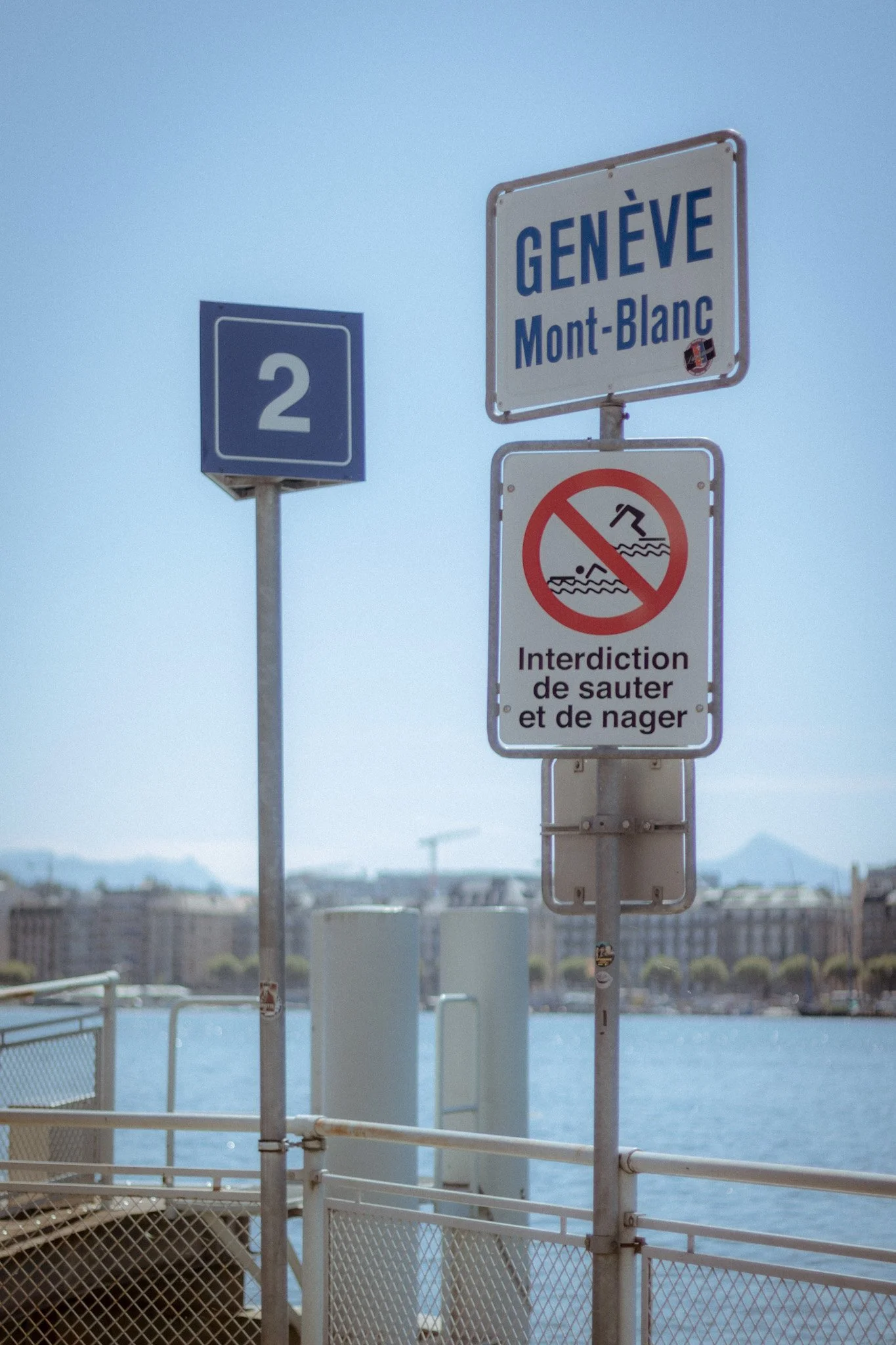 Signs along a waterfront, including a blue highway sign with the number 2, a directional sign for Geneva and Mont-Blanc, and a no swimming or diving warning in French.