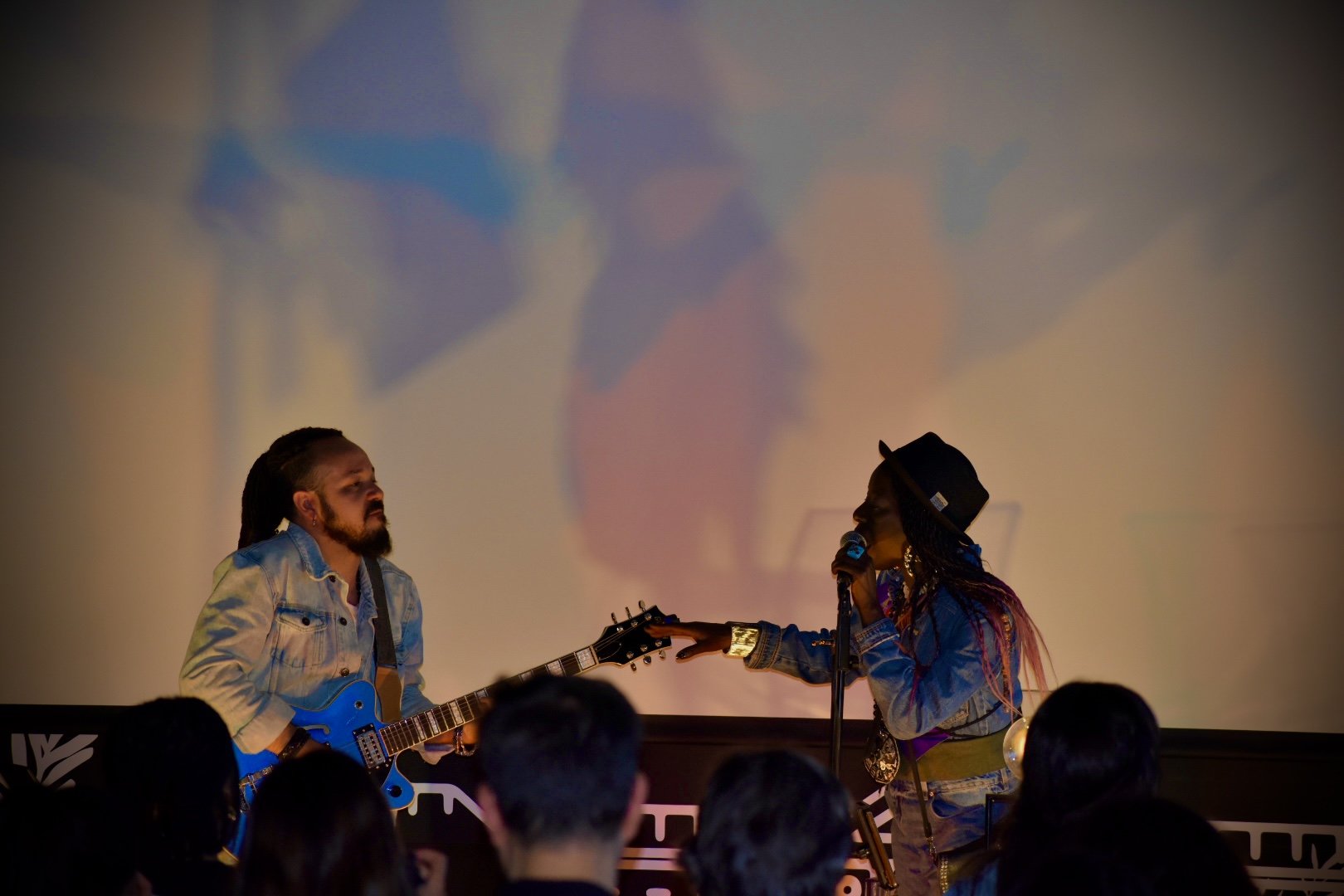 Two musicians perform on stage illuminated by colorful light. One is a man with long dreadlocks playing an electric guitar, and the other is a woman with long braids, a hat, and a denim jacket, singing into a microphone.