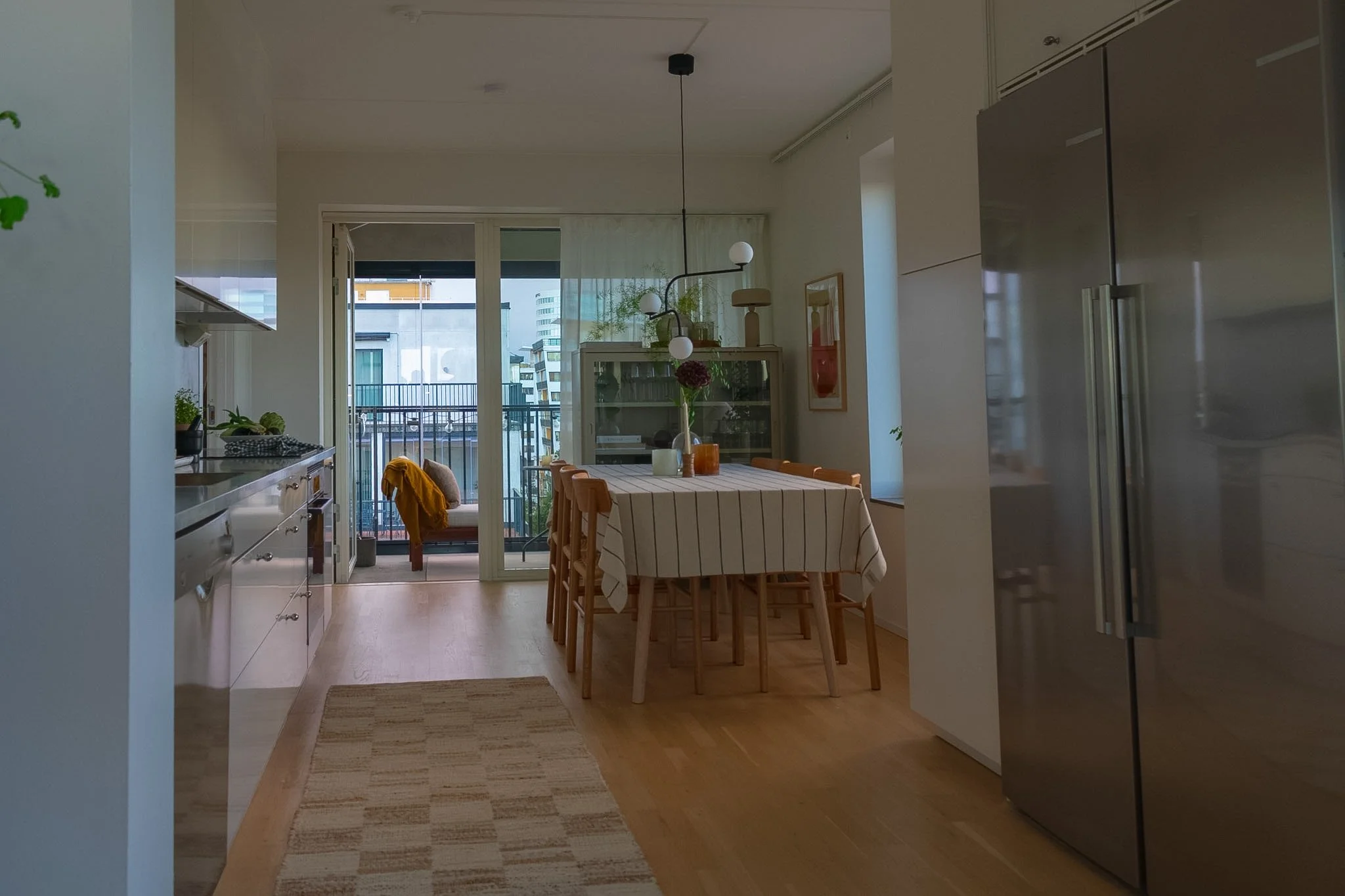 Interior of a modern apartment with a dining area near a balcony, decorated with plants and artwork, featuring a wooden dining table with chairs, a white cabinet, and a large stainless steel refrigerator.