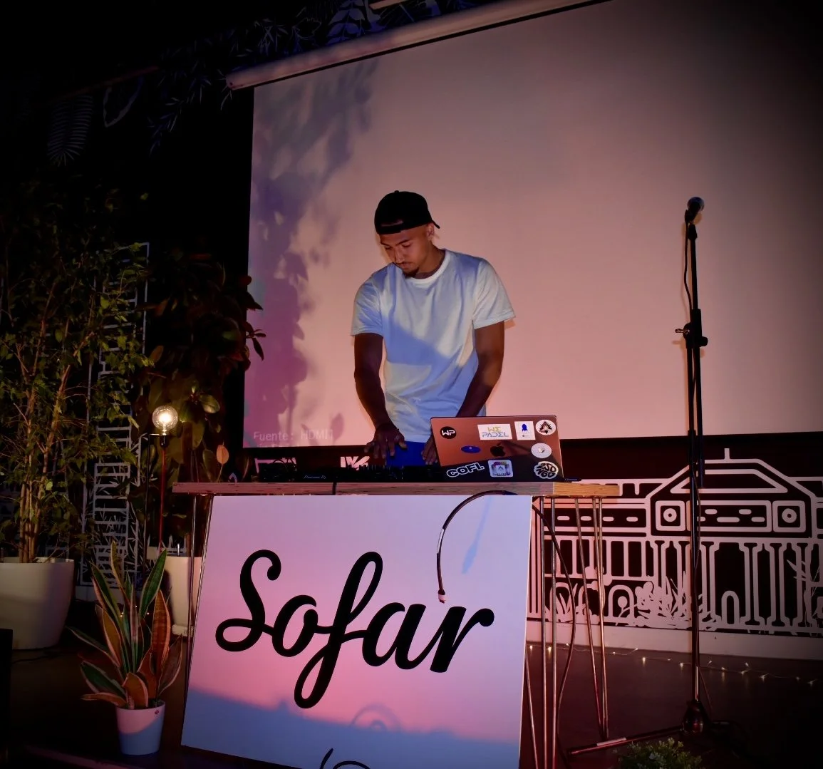 A DJ performs at a pink-themed event called SoFar, standing behind a table with DJ equipment, a laptop, and stickers. The background has a large screen with a purple and pink gradient, and there are potted plants and a microphone on a stand nearby.