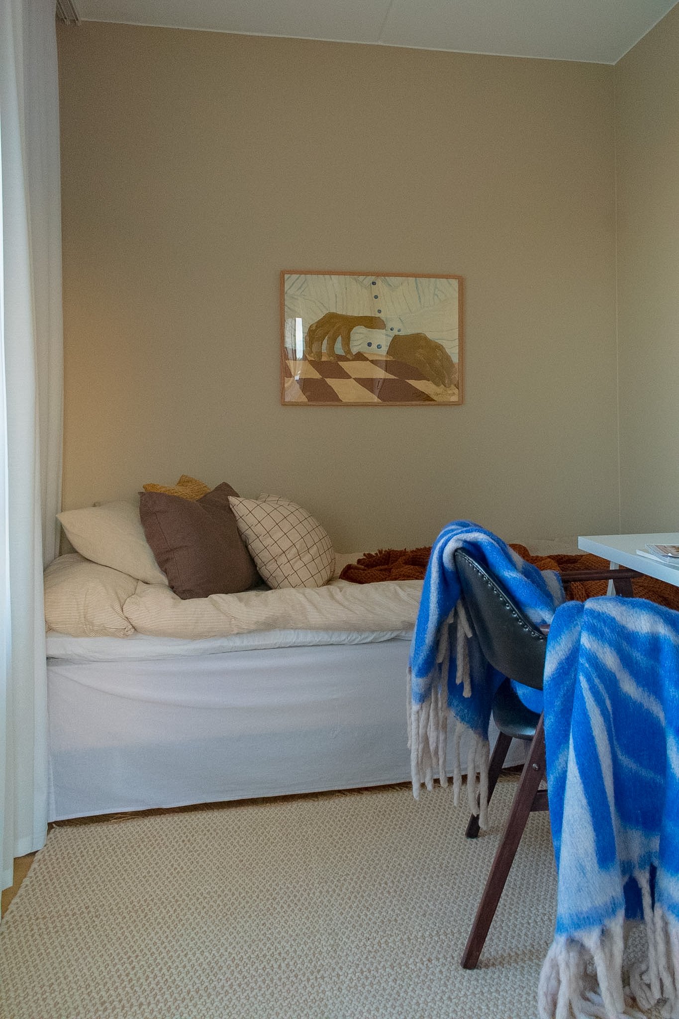A bedroom with a bed on the left side, decorated with pillows and a beige blanket. A framed picture of a dog with a collar is hanging on the plain beige wall. There is a table with clothes or towels draped over a chair on the right side, with a blue 