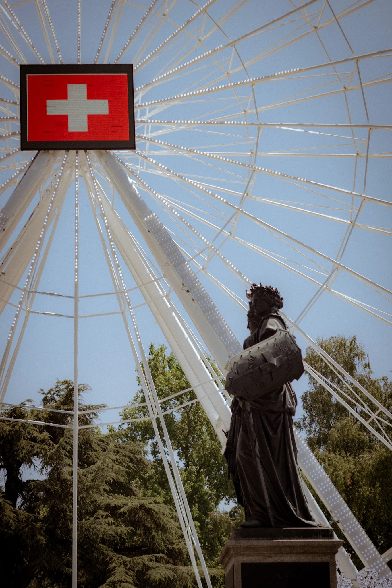 Looking up at a large Ferris wheel with a red Swiss flag sign at the top, against a blue sky.