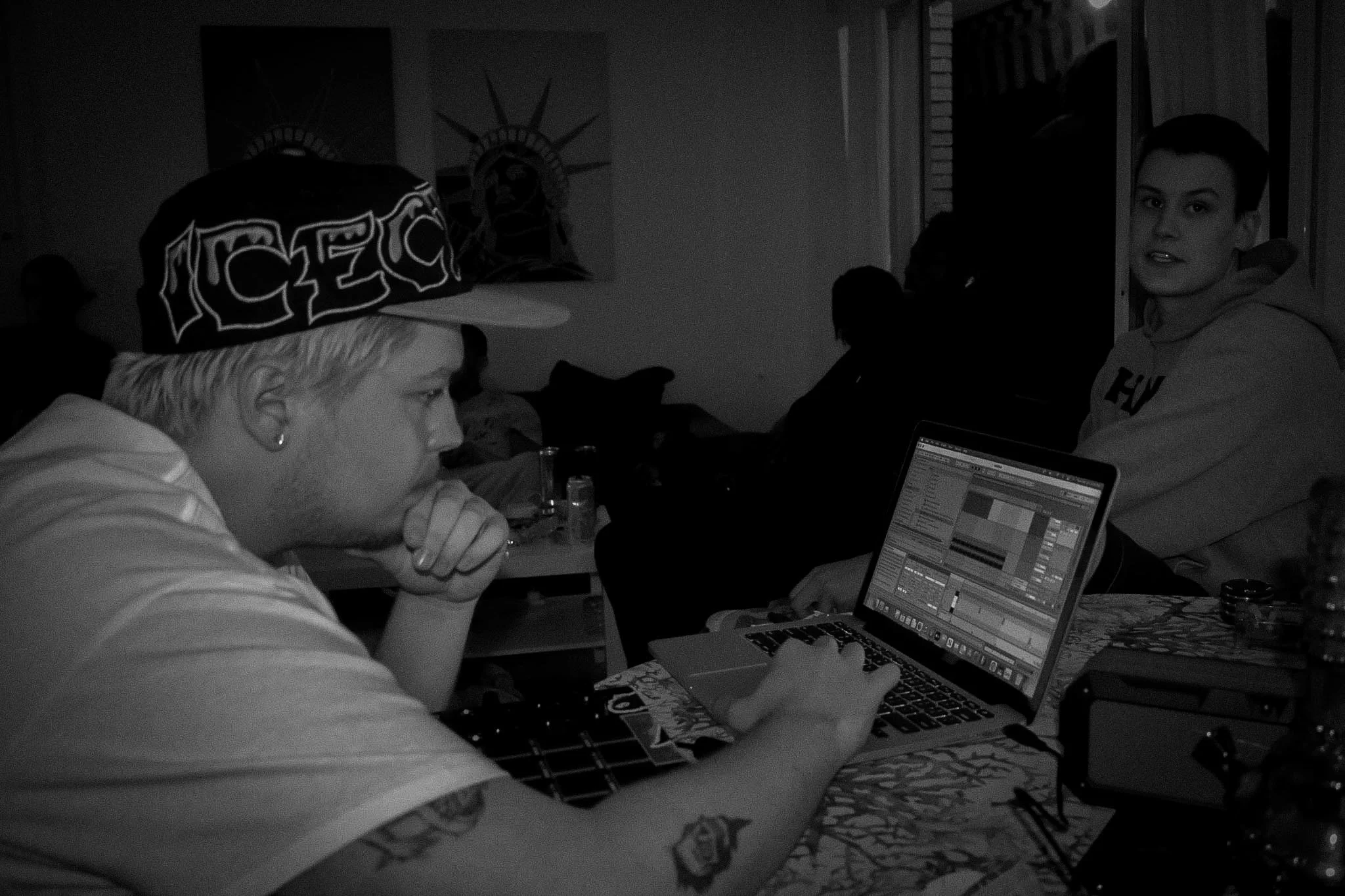 A woman with short hair sitting at a desk with a laptop, surrounded by people, in a dimly lit room. Art on the wall and a person with a hat in the foreground.