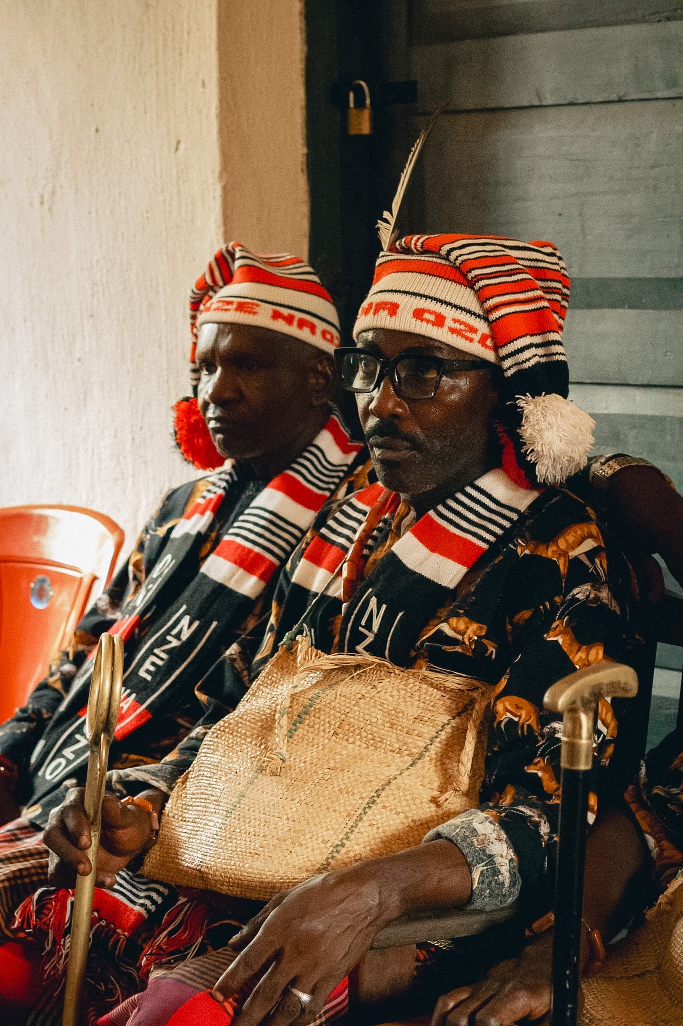 Two men in colorful striped traditional African attire and headwear sitting side by side, one with glasses and a walking stick, in an indoor setting.