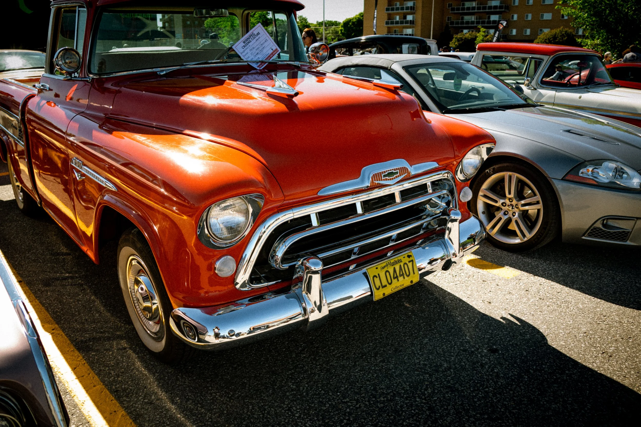 A red vintage Chevrolet truck parked in a lot at a car show, with other classic and modern cars in the background.