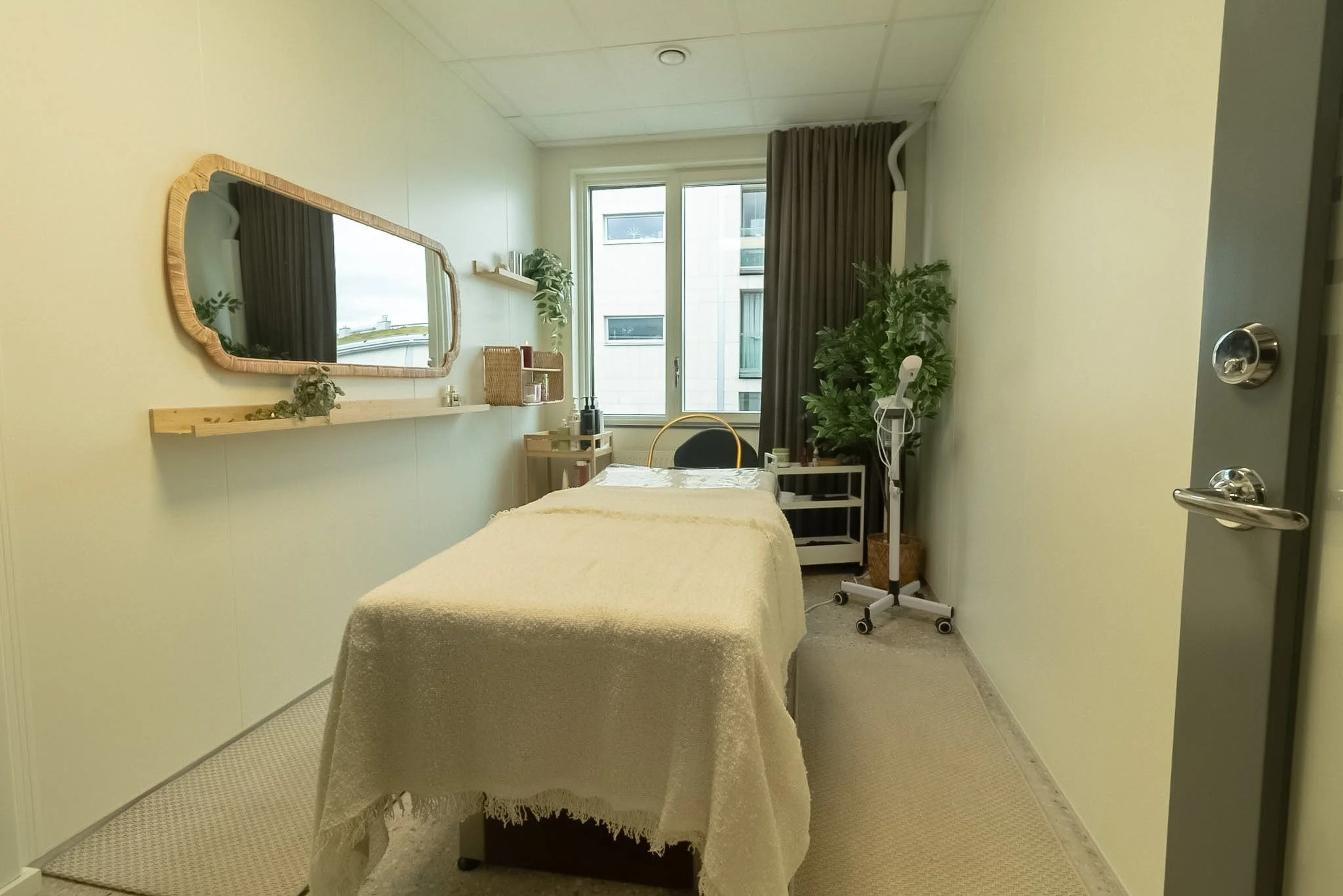 A small room with a massage table covered in a beige blanket, a large window with dark curtains, a mirror on the wall, and various plants and storage shelves.