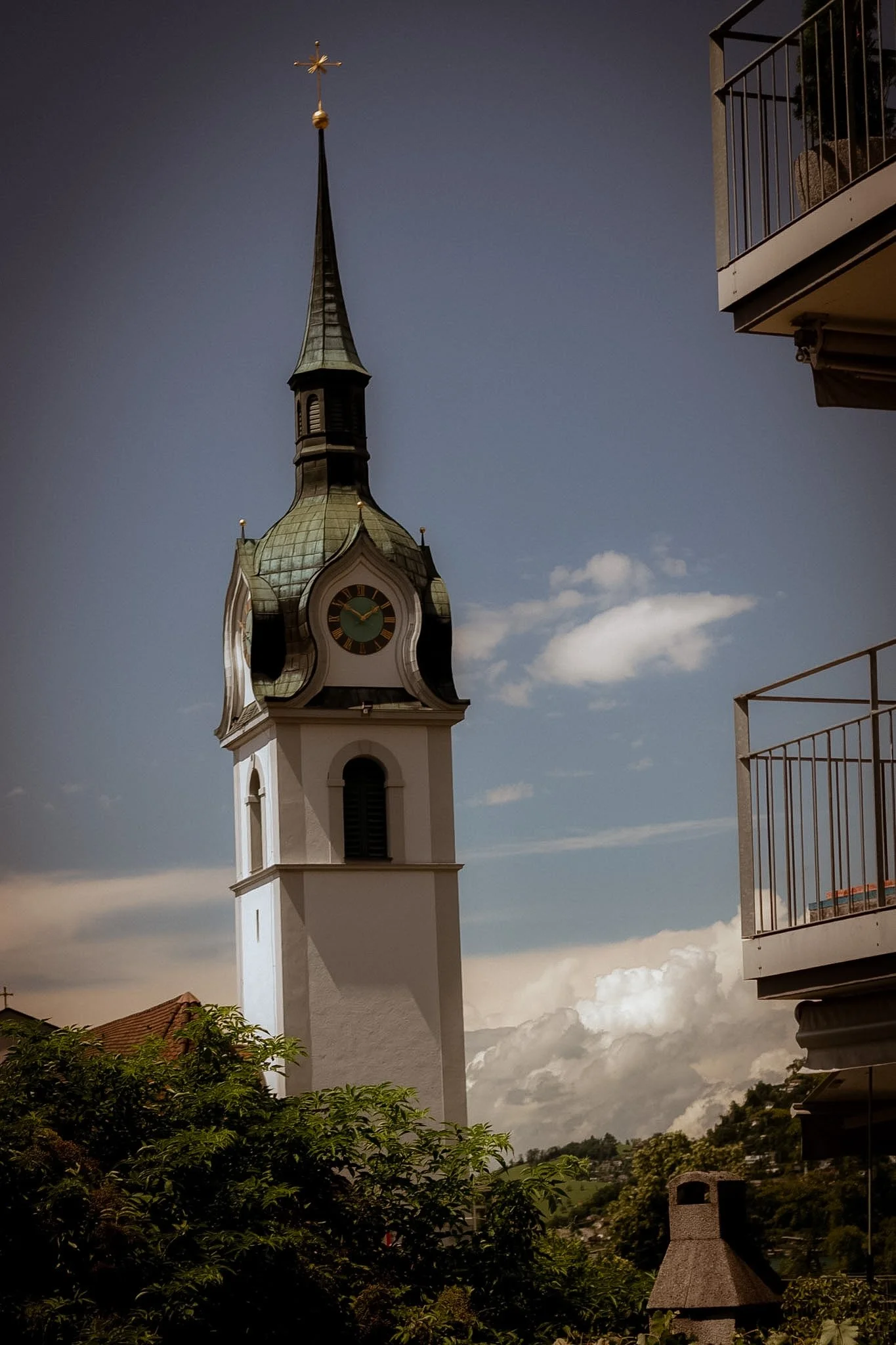View of a church steeple with a clock, a golden cross on top, and a cloudy sky in the background, with part of a building balcony on the right and trees at the bottom.