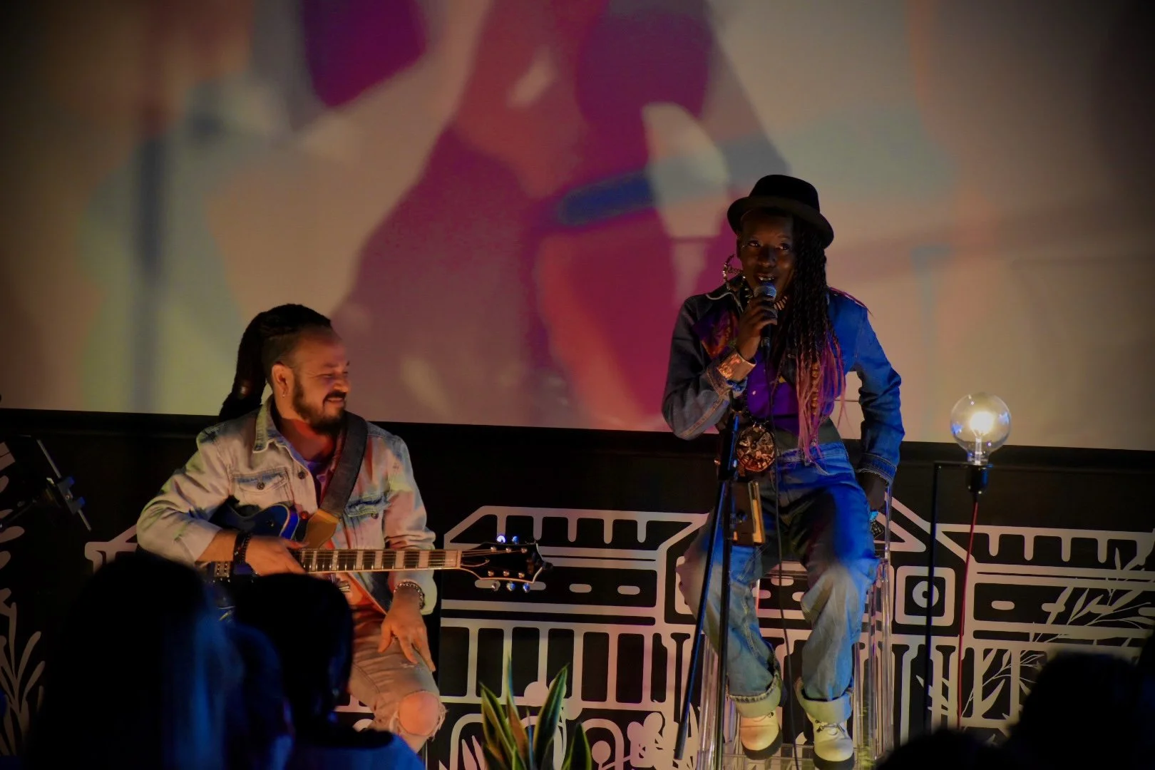 Two musicians performing on a stage with a large screen displaying their image in the background. One is sitting and playing a guitar, while the other is standing and singing into a microphone. The stage has a black and white patterned design.