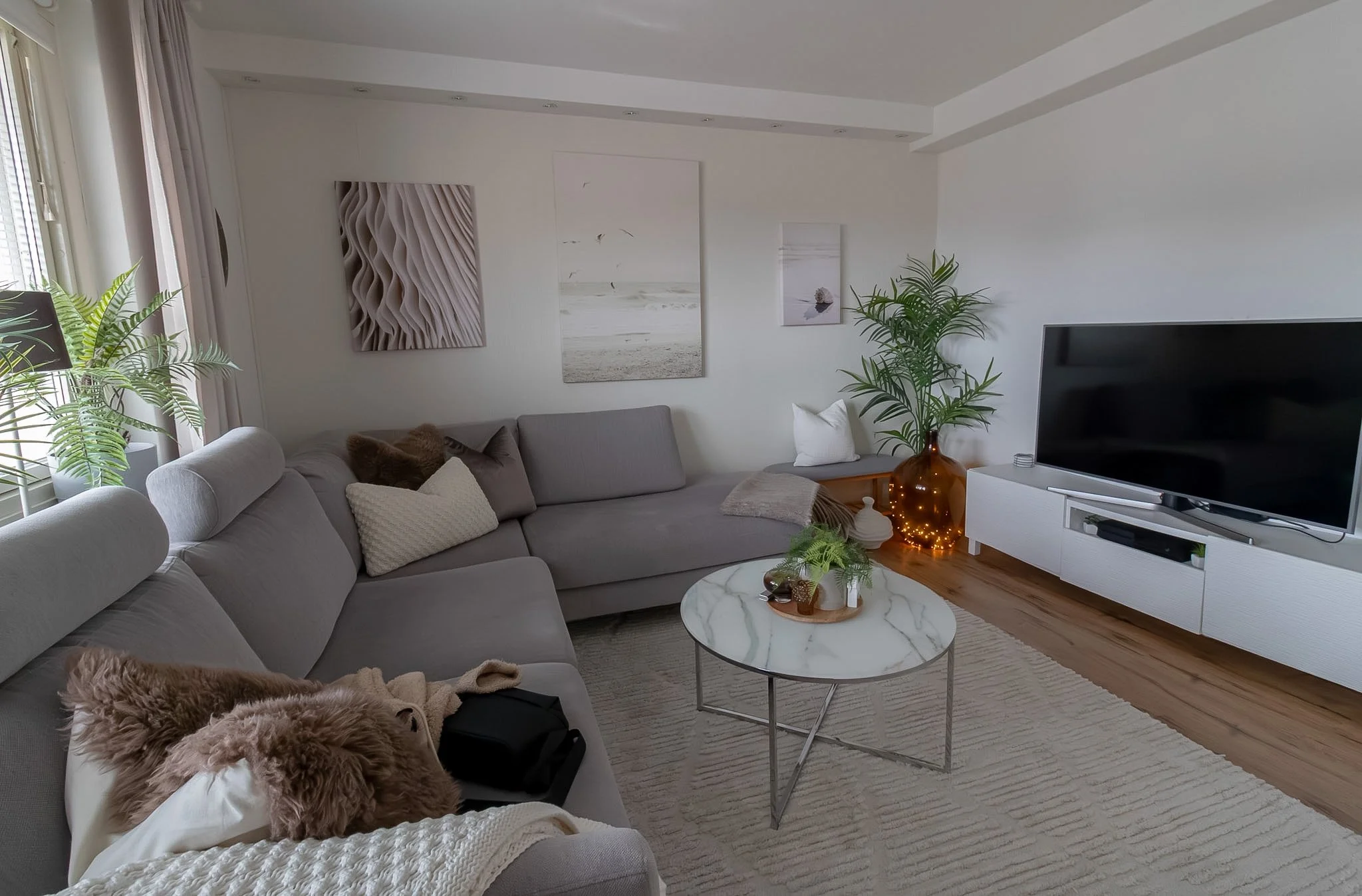 A living room with a gray couch decorated with pillows and a fur blanket, a round marble coffee table, a flat-screen TV on a white cabinet, potted plants, wall art, and a large window with curtains.