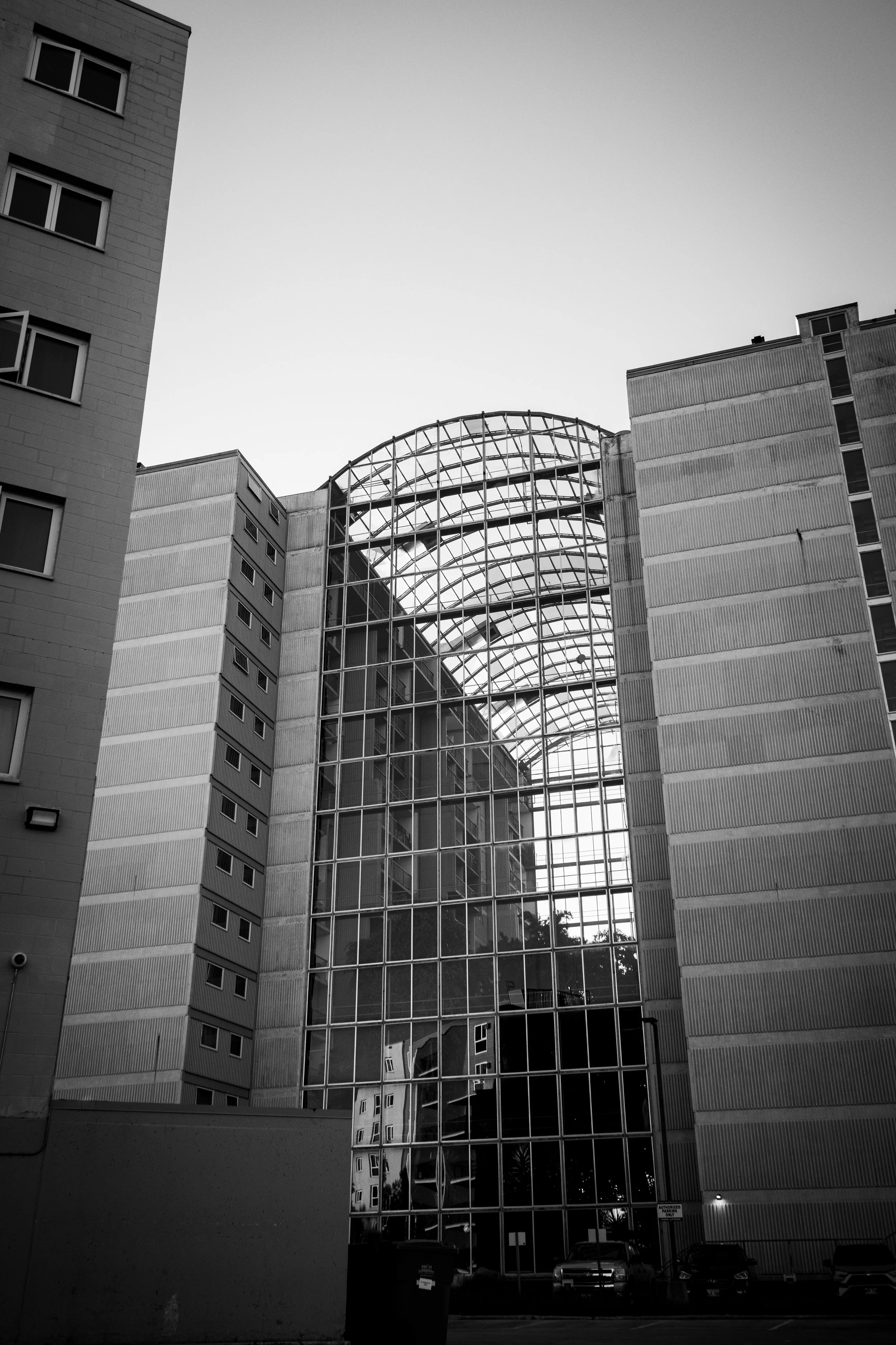 Black and white photograph of a modern building with a large curved glass and metal atrium between two taller sections of the building. Several cars are parked in front of the building.