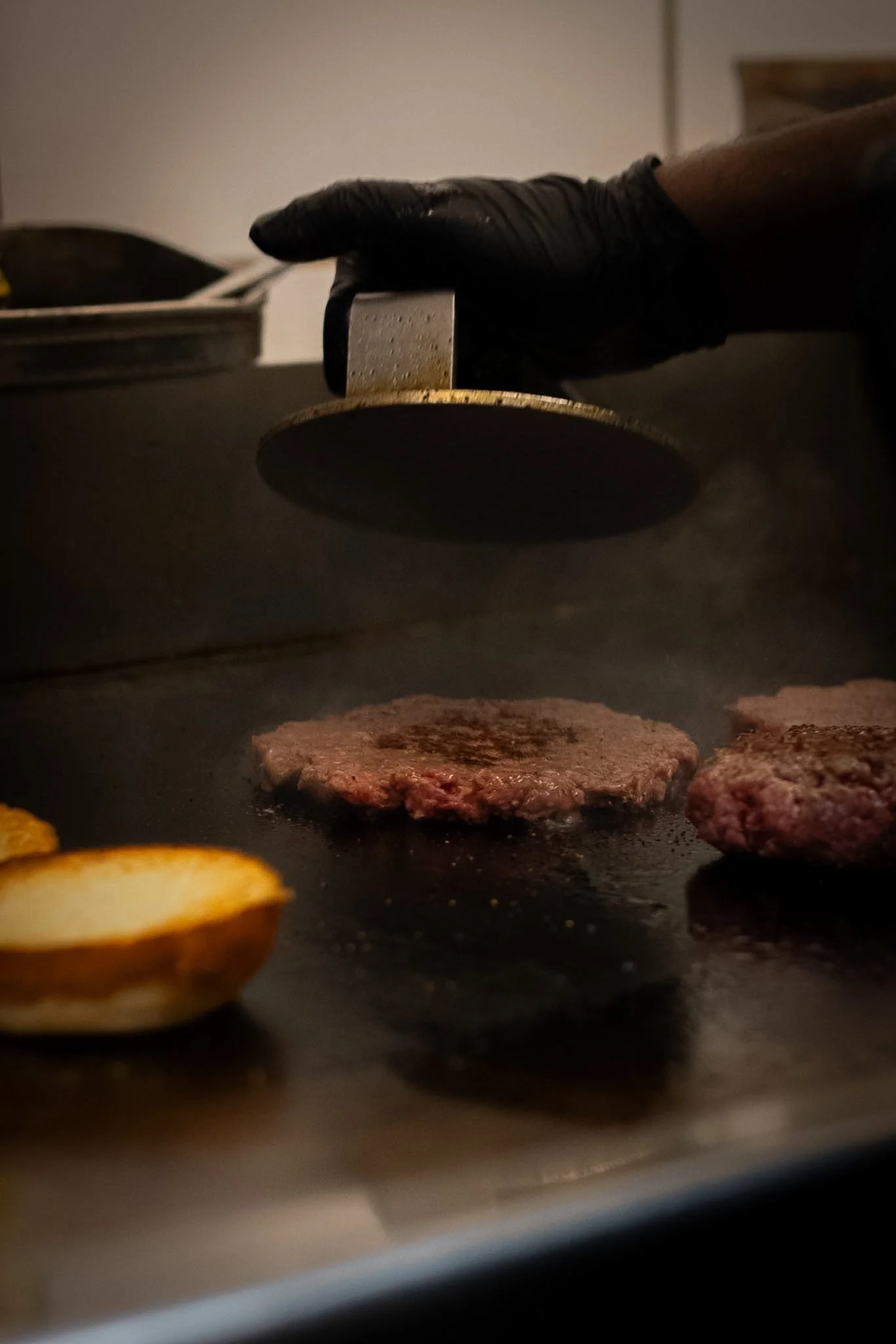 A person using a meat tenderizer on a hamburger patty on a griddle. Other food items are visible nearby.
