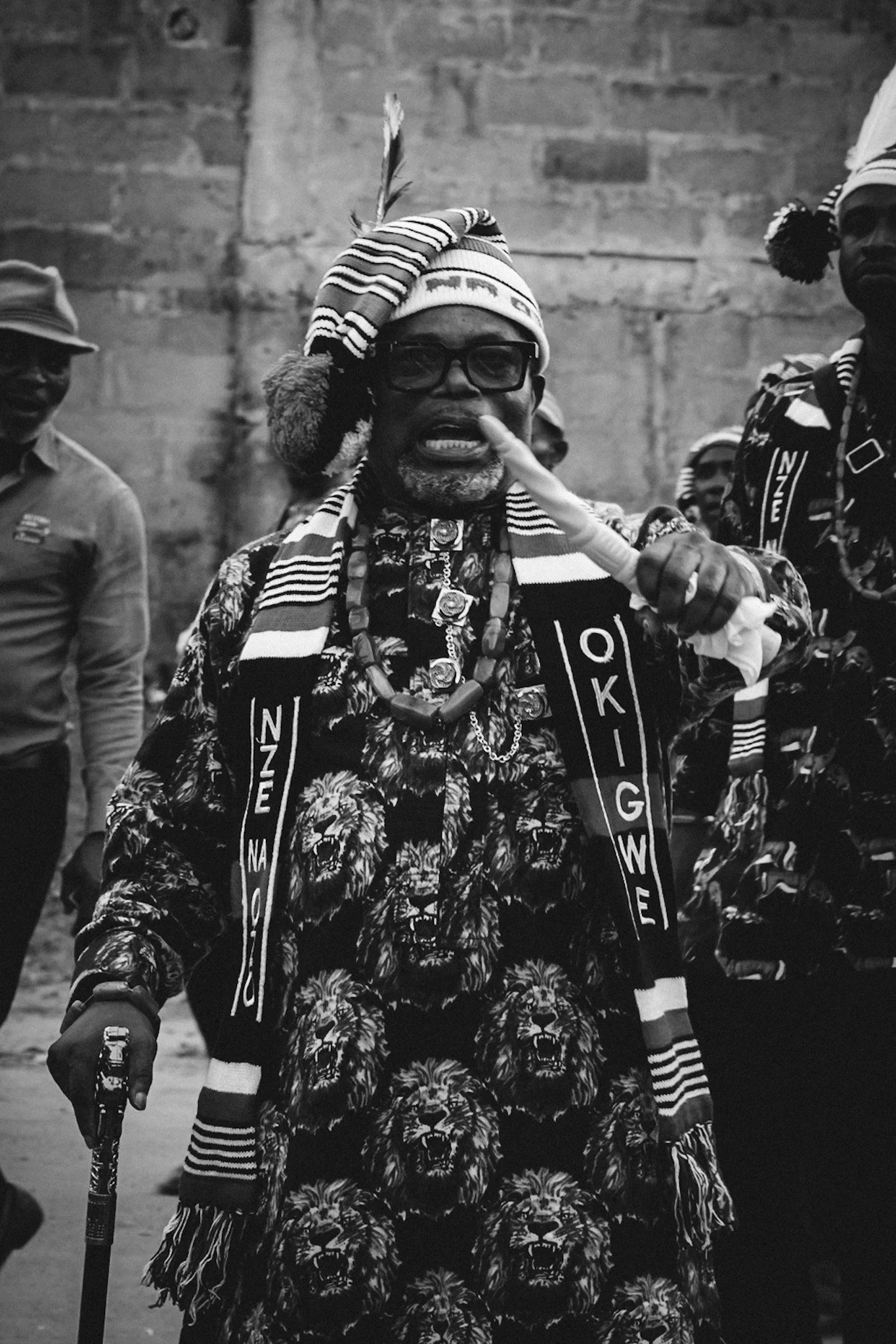 A man wearing a patterned shirt and multiple necklaces, with a striped scarf and a beanie with a feather, making a gesture with his index finger. In the background, there are other people and a brick wall.