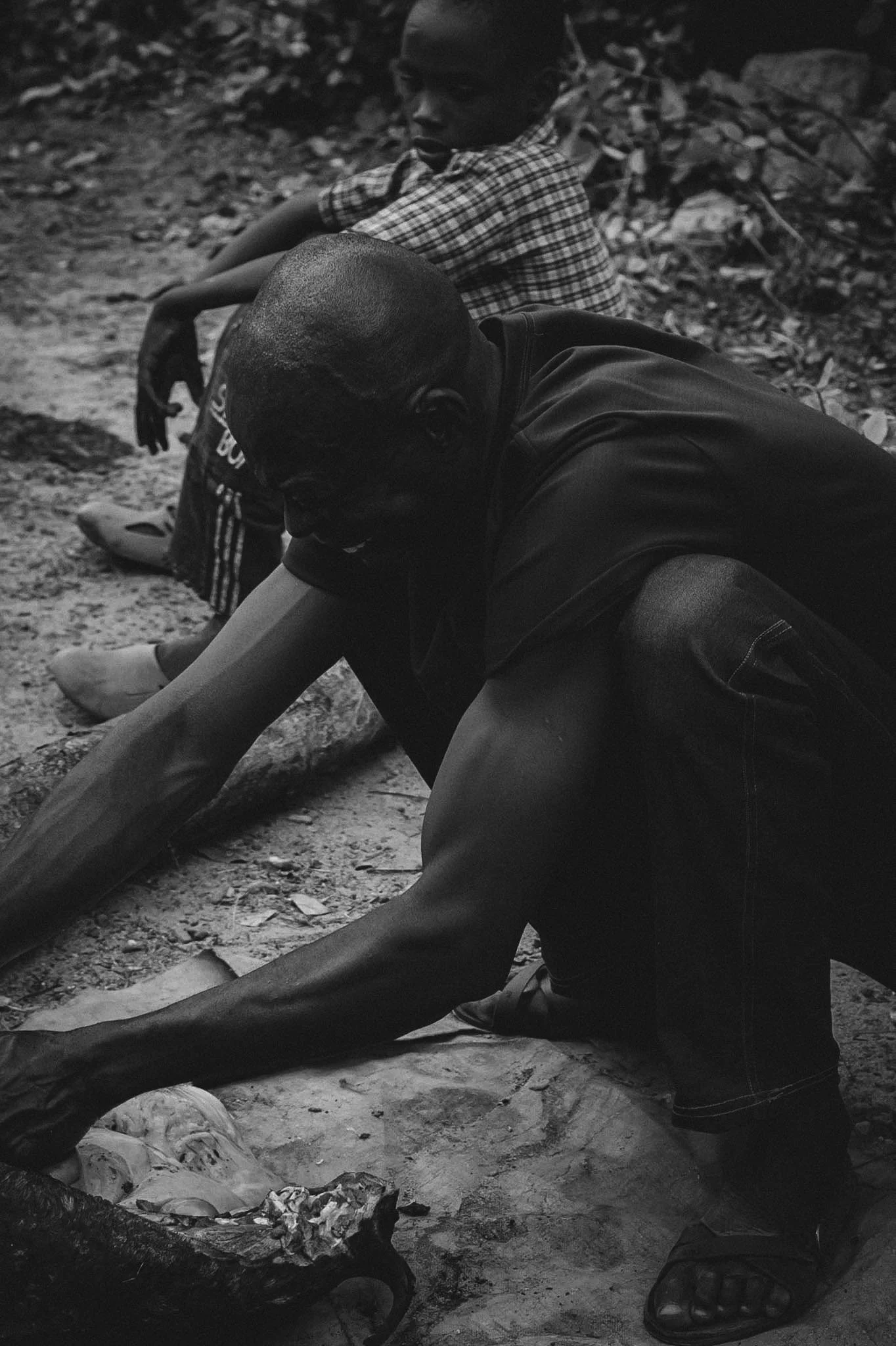Two people sitting on the ground outdoors, one wearing glasses, a checkered shirt, and sandals, the other wearing a dark shirt, shorts, and sandals, with a foot resting on a piece of a carved or sculpted object.
