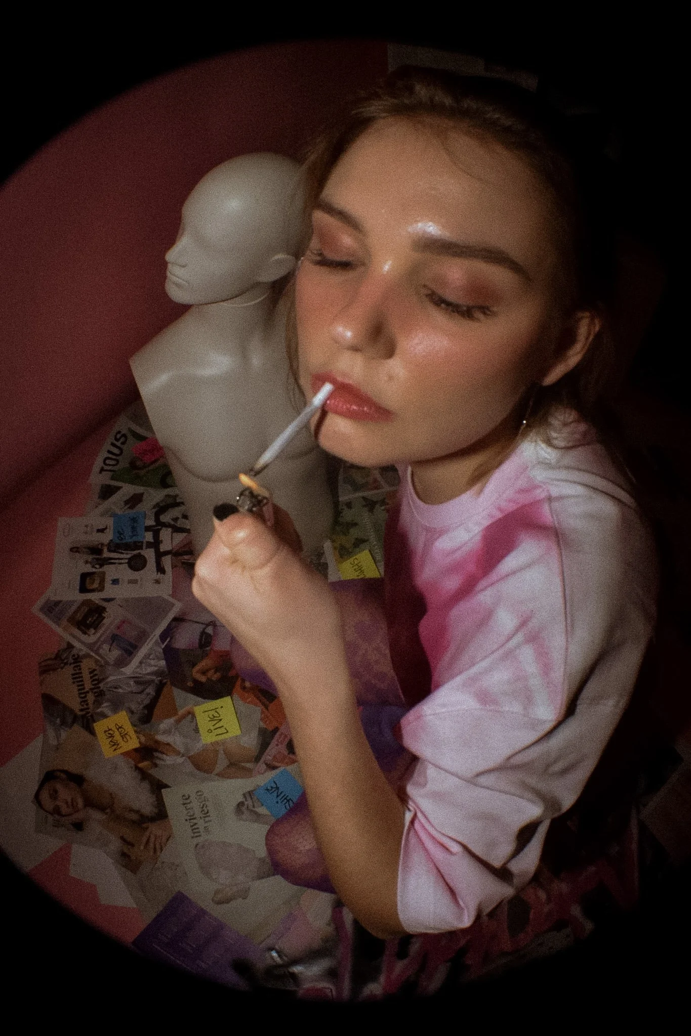 A young woman with light brown hair and makeup, wearing a pink and white tie-dye shirt, is lighting a cigarette or joint. She is positioned beside a white mannequin head with closed eyes. The background features a pink wall and a bulletin board with 