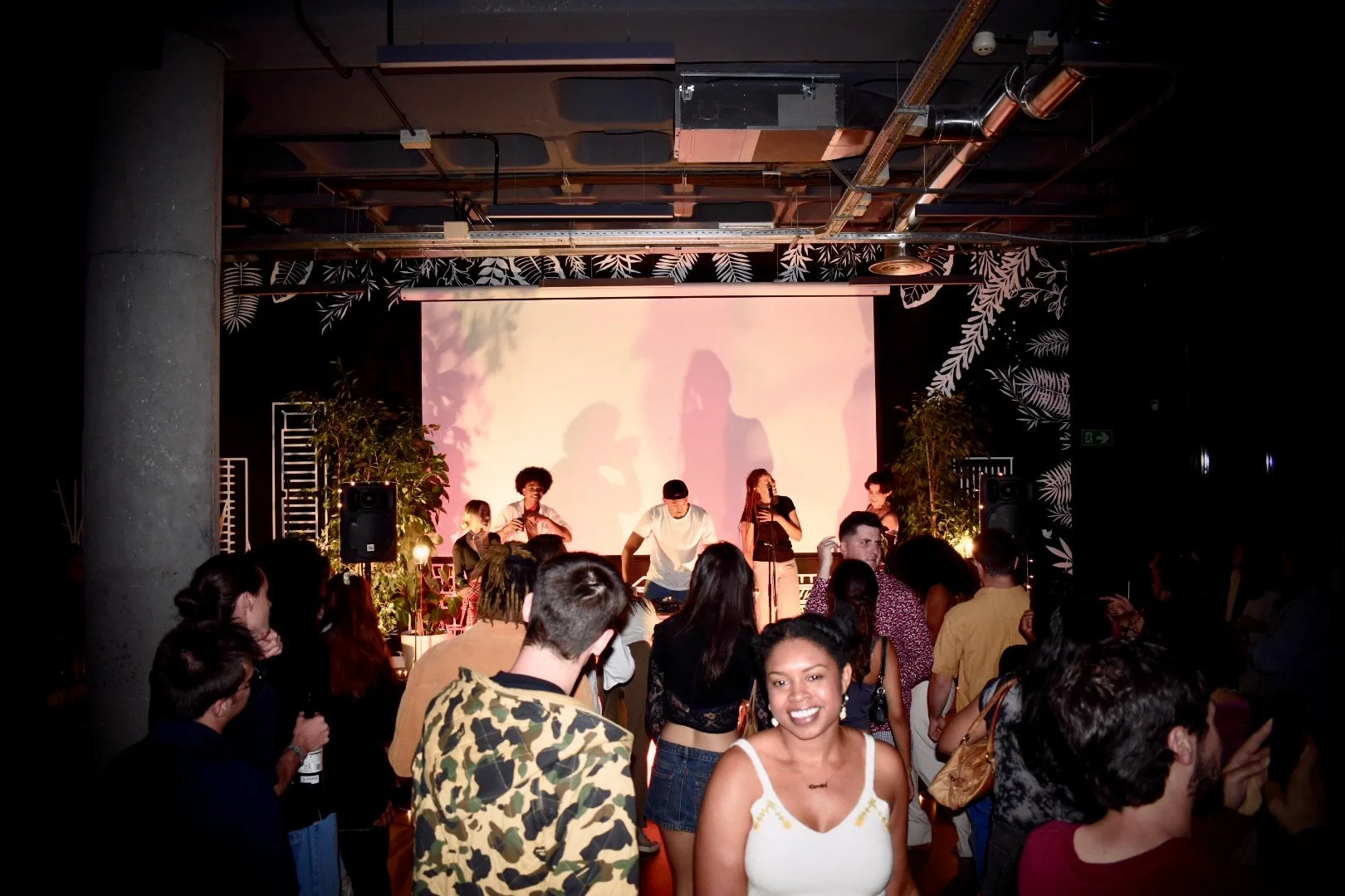 A diverse crowd of people at a live music event in a dimly lit venue with a large screen and tropical-themed wall decorations.