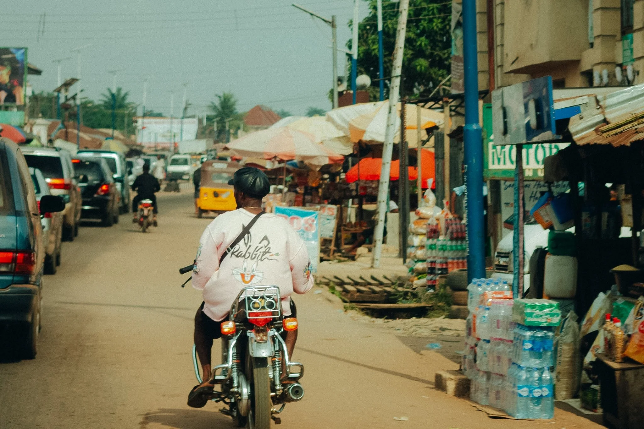 A person riding a bicycle down a busy street with small shops and stalls on the sides, cars and motorcycles on the road, and umbrellas covering market stalls, with palm trees and buildings in the background.