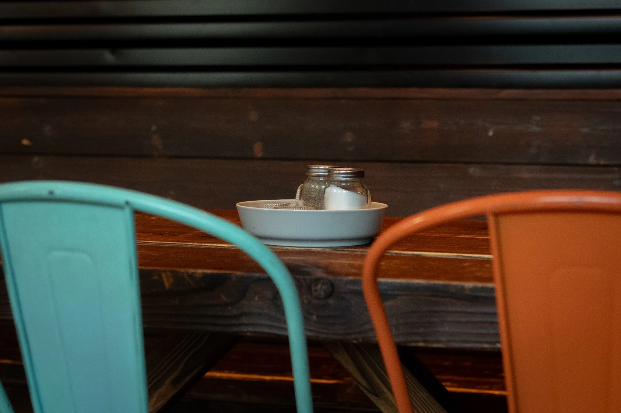 A table at a restaurant with three salt and pepper shakers in a white bowl, and three chairs in front of it, two orange and one turquoise.