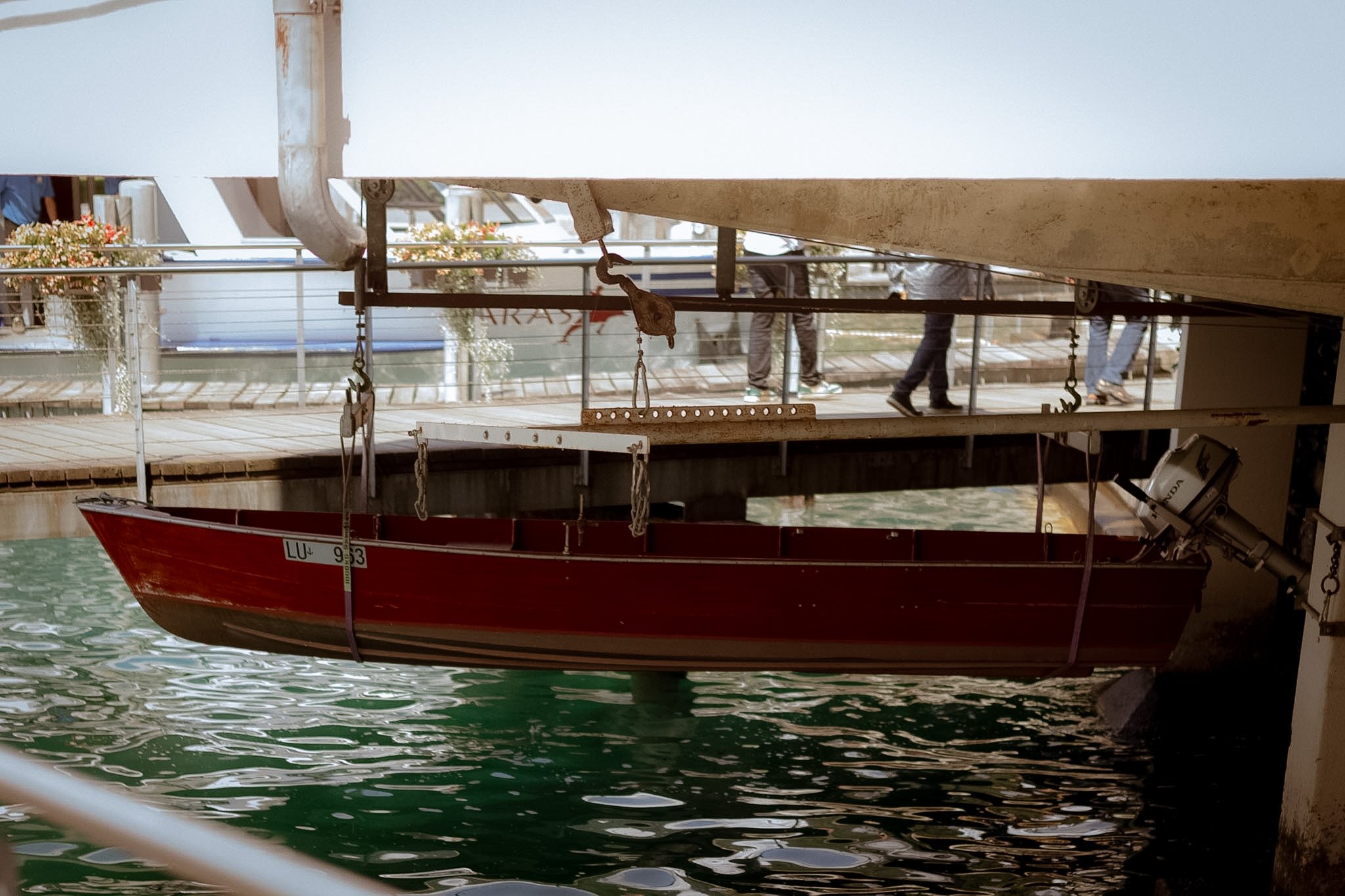 A red boat is docked under a bridge, with water reflecting the boat and the surrounding structures. Part of the bridge and a few people walking on a nearby walkway are visible.