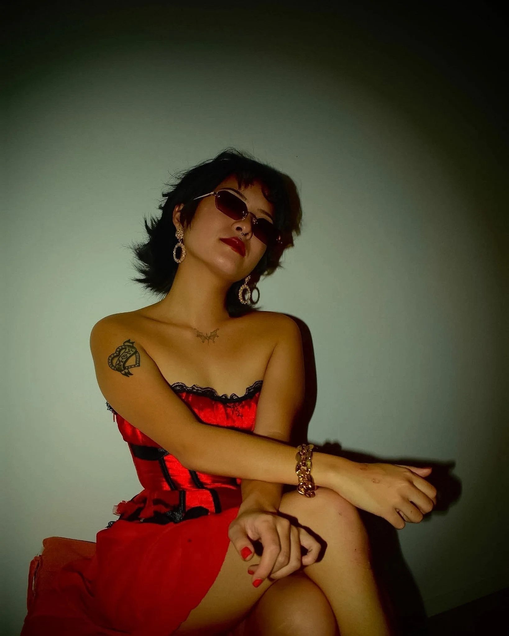 A woman with short black hair, wearing sunglasses, red lipstick, and gold jewelry, is seated and partially dressed in lingerie, with tattoos visible on her arm.