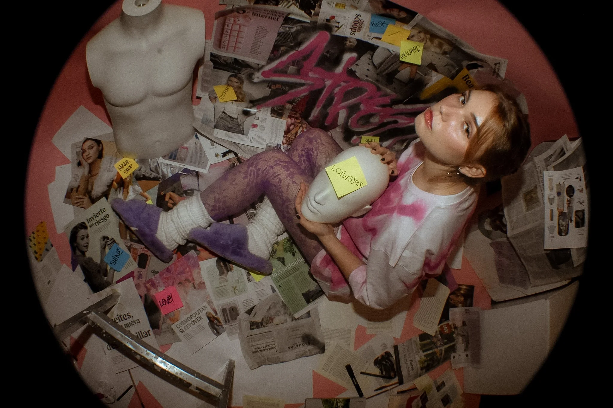 A woman with reddish-brown hair and makeup, wearing a pink tie-dye shirt, lies amid scattered magazines, photos, sticky notes, and a white mannequin head with a label reading "LOUISER."