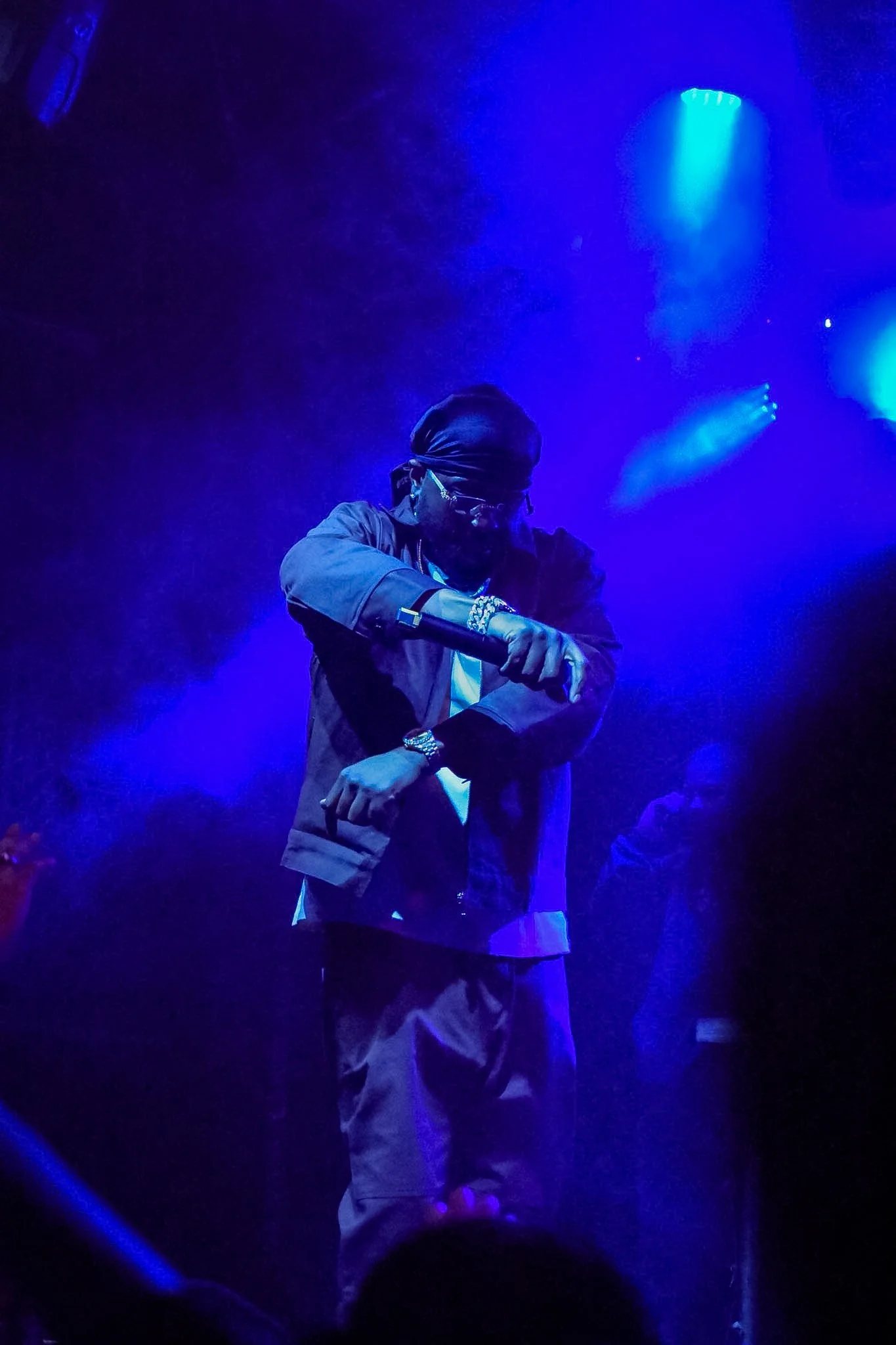A person dressed in a black jacket, beige pants, wearing jewelry, sunglasses, a black head covering, and gloves, dancing or performing under blue stage lighting in a dark environment.