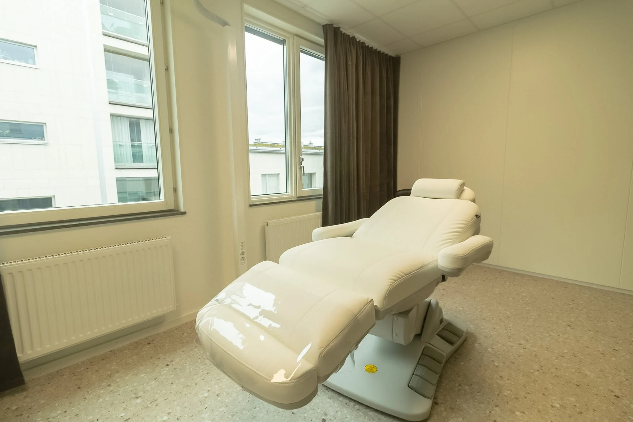 Medical examination room with a white reclining medical chair near large windows with brown curtains, overlooking neighboring buildings.