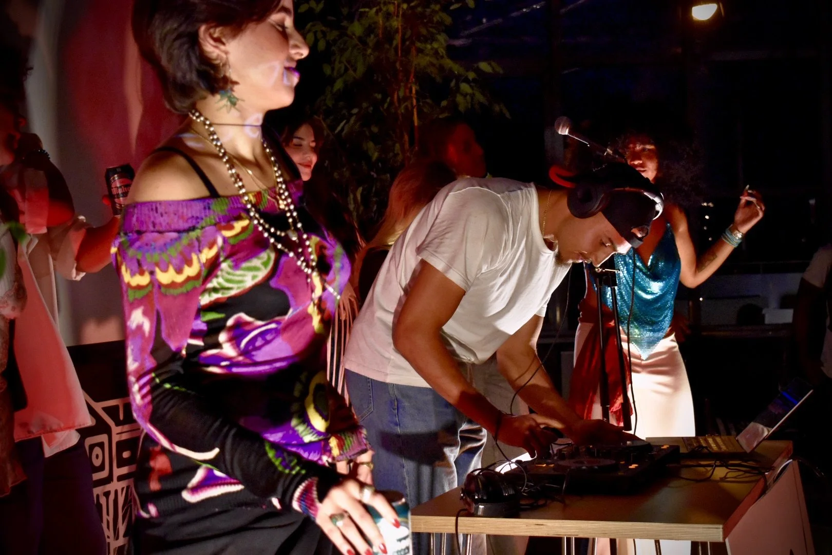 People dancing and DJ in a dimly lit setting, some with headphones, surrounded by colorful outfits and musical equipment.