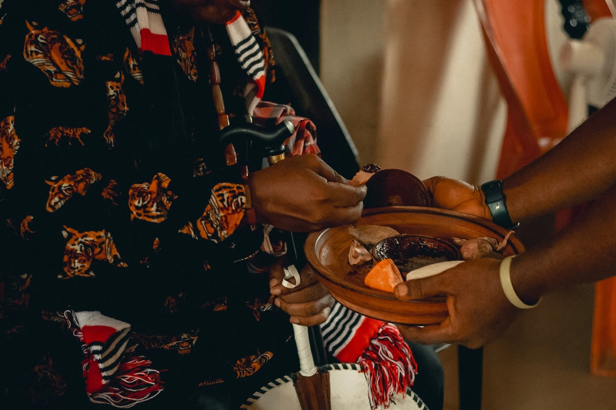 A person wearing a black patterned shirt with orange tiger designs and red, black, and white accessories is holding a wooden platter filled with grilled meats, vegetables, and sauces. The person's left hand, which has a dark ring, is resting on the p