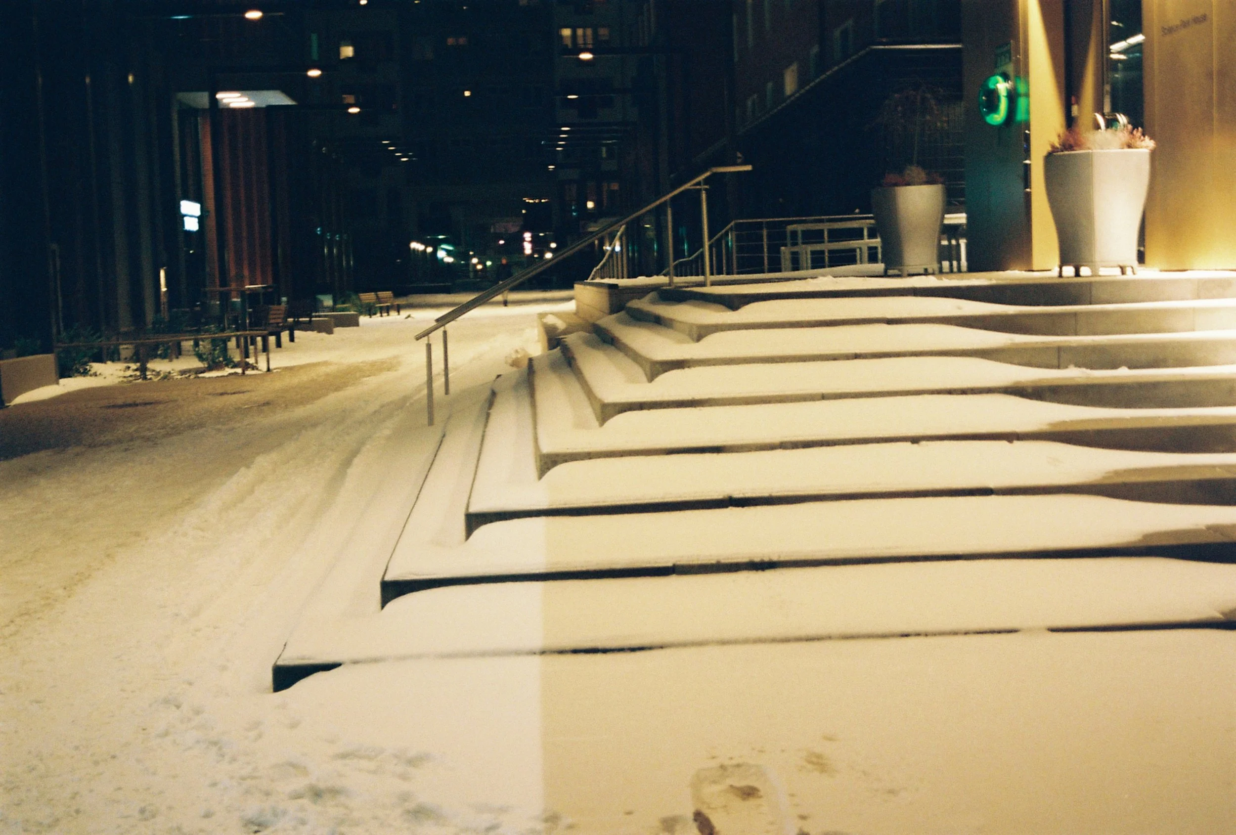 Snow-covered outdoor stairs and sidewalk at night, with large potted plants, yellow wall, and illuminated building in the background.