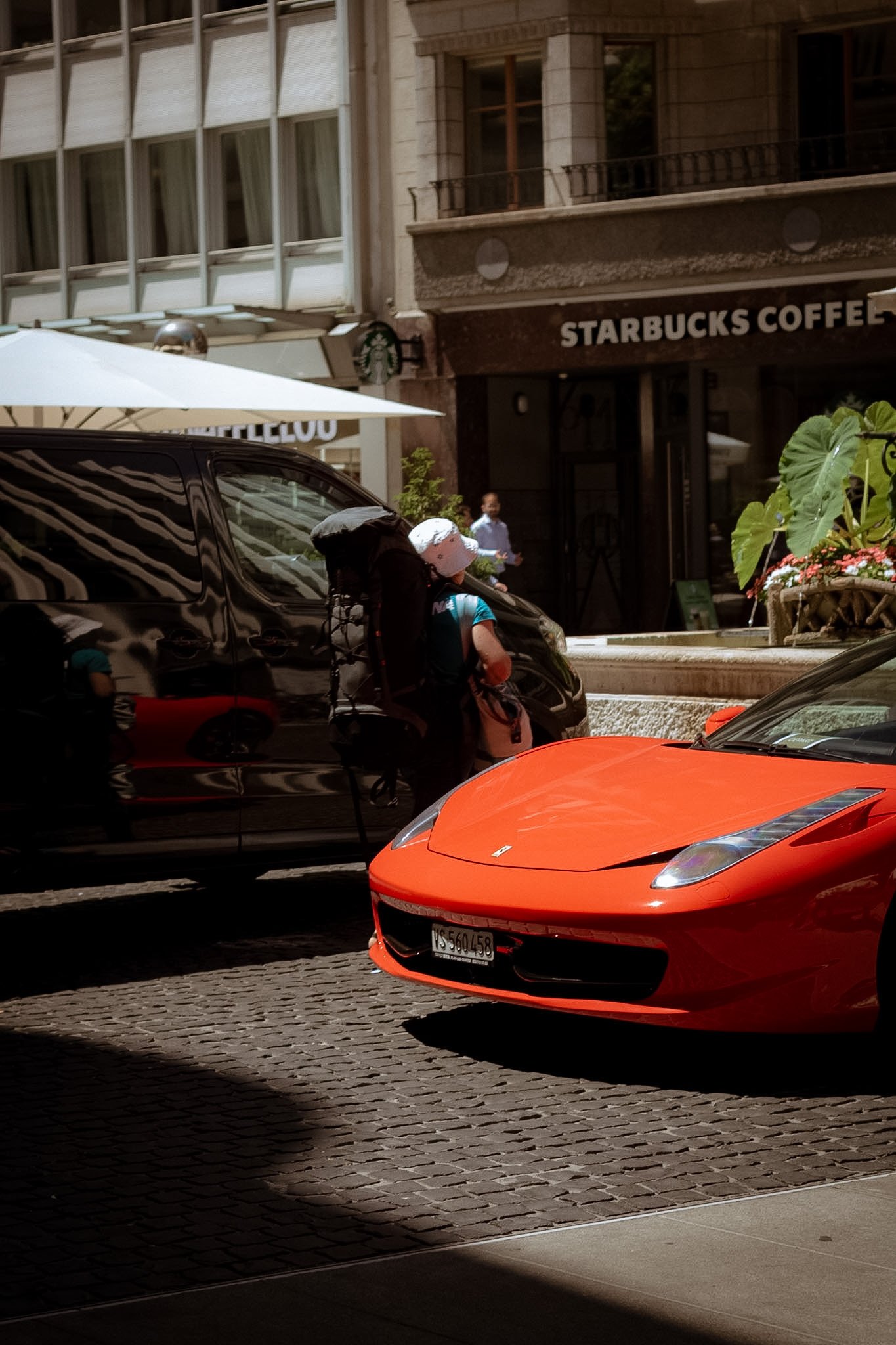 Street scene with a bright orange sports car, black car, people walking, and a Starbucks Coffee shop in an urban area.
