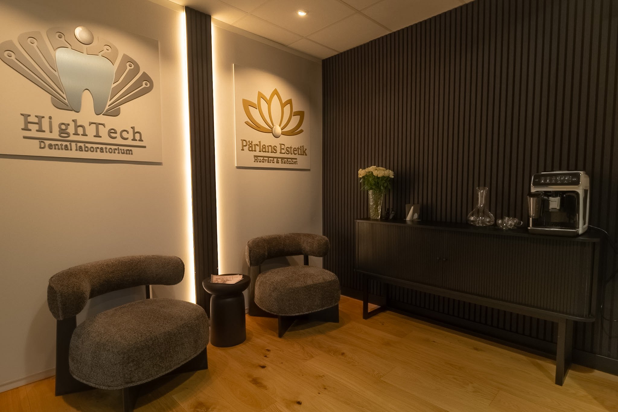 Interior of a waiting area with three upholstered chairs, a small round table, a black cabinet with a vase of white flowers, a coffee machine, and wall signs for HiGHTech Dental Laboratory and Pärlans Estetik.
