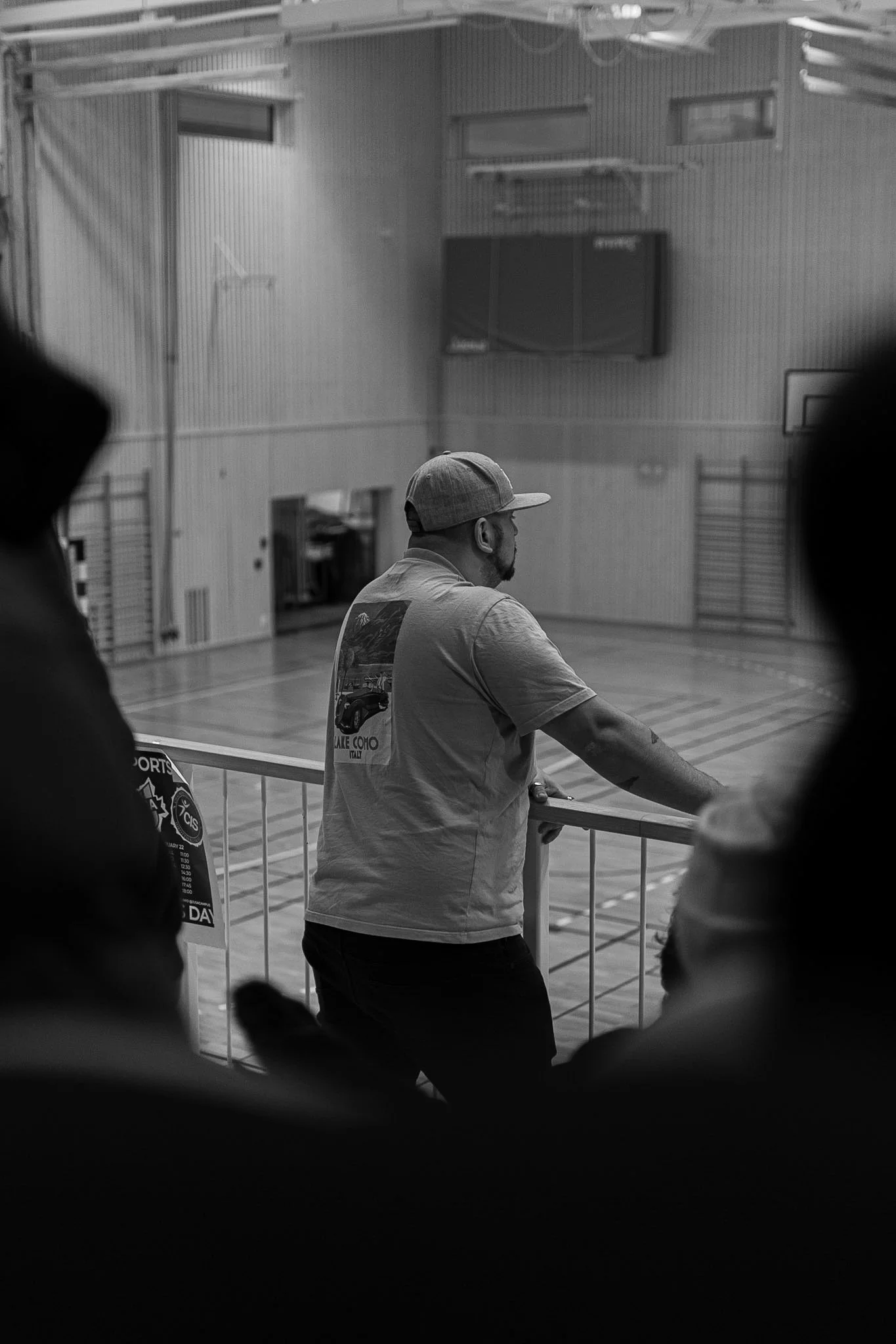 A man in a baseball cap and T-shirt stands in a gymnasium, leaning on a railing, with other individuals in the foreground and background, some partially visible. The gym has a basketball hoop, wall-mounted TV, and gymnastic equipment.