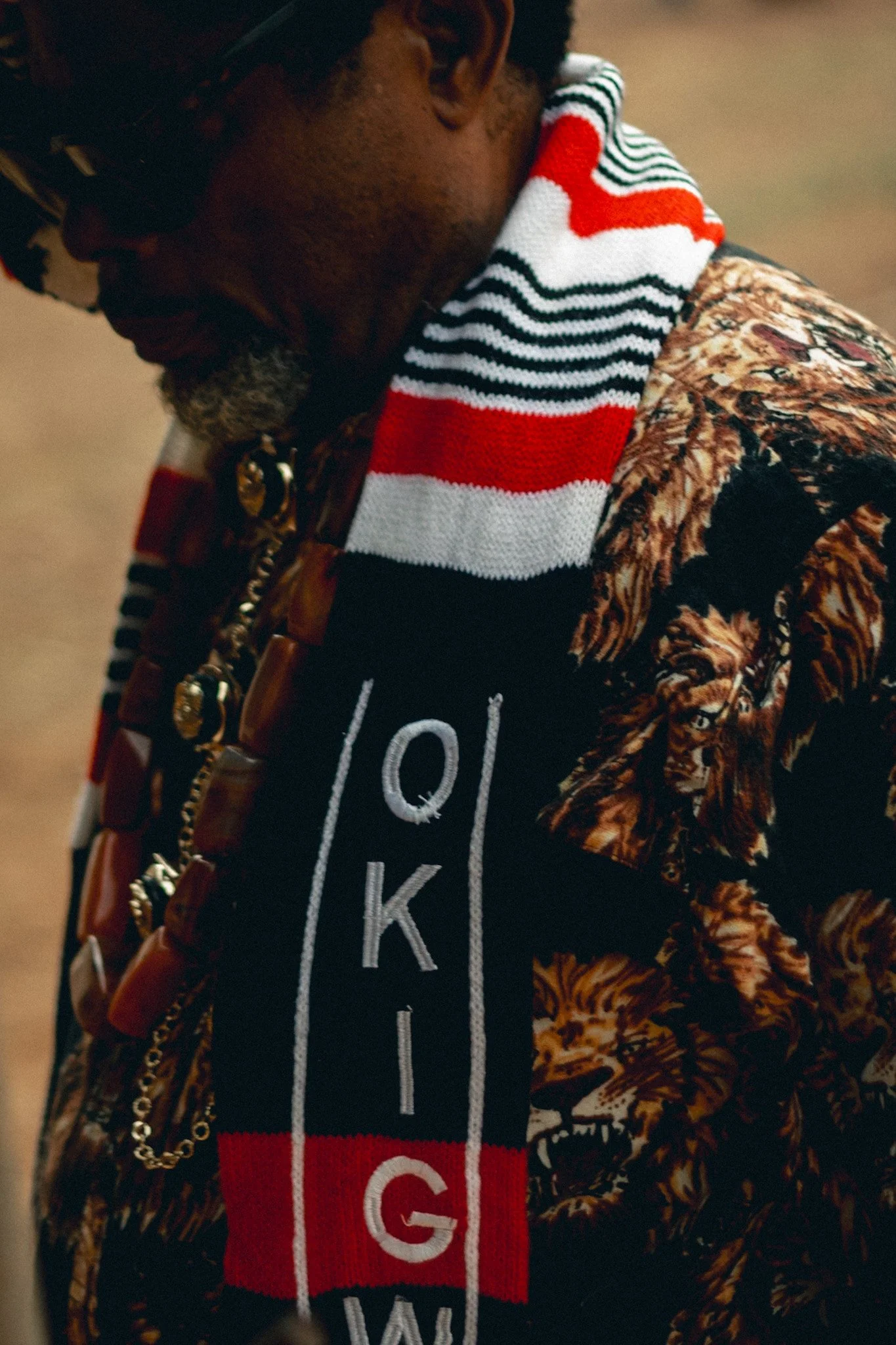 A person is resting their head on their hand, wearing a patterned jacket and a scarf with red, white, and black stripes that reads 'GLOW' and has a red heart.
