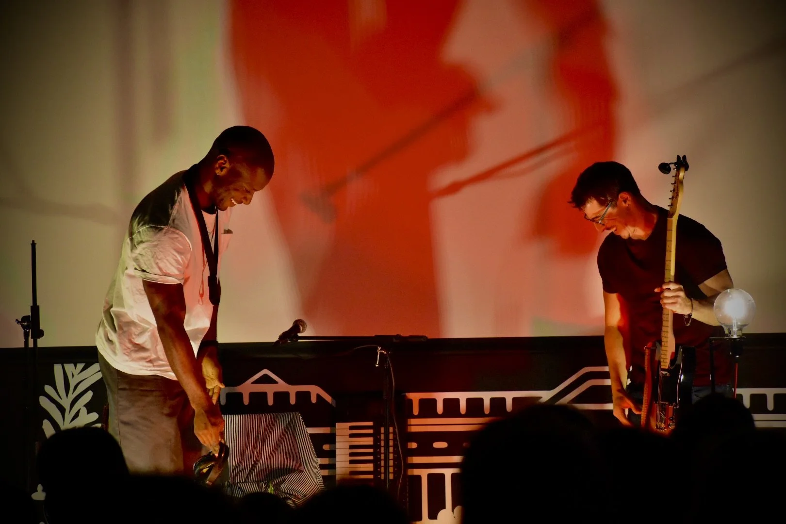 Two musicians bowing on stage, one with headphones and a keyboard, the other holding a guitar, with a red and orange abstract background projection.