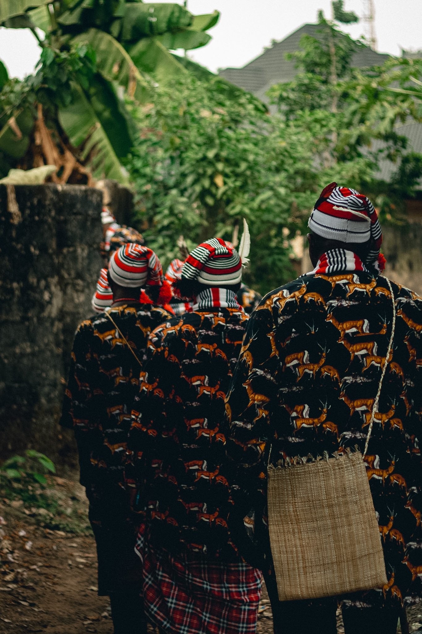 People walking outdoors wearing colorful, patterned clothing and striped hats, carrying a woven bag, with greenery and a house in the background.