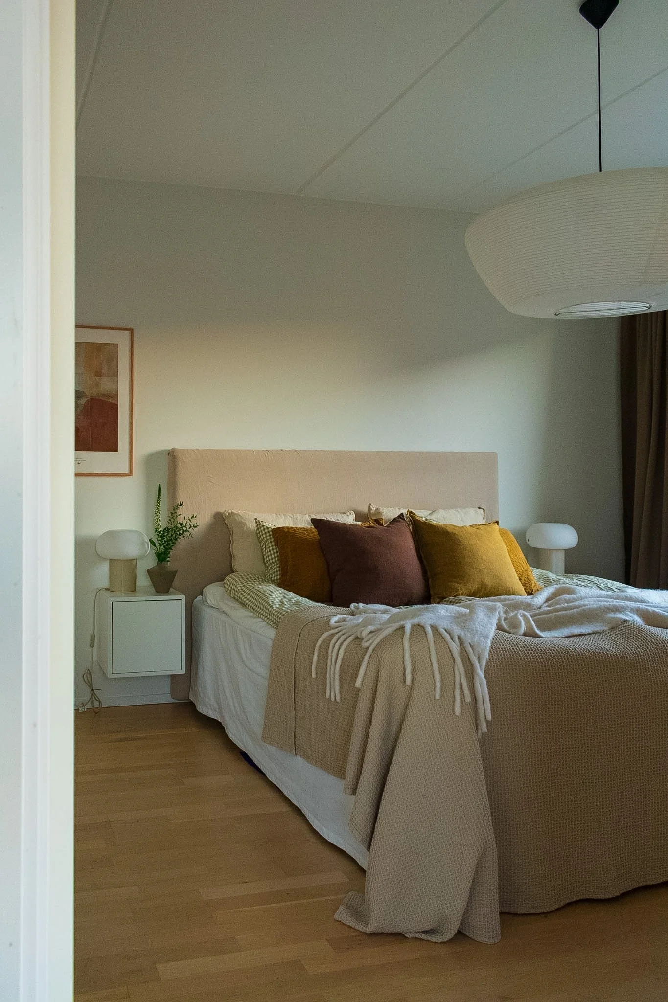 A cozy bedroom with a large bed covered with beige and mustard-colored pillows, a cream blanket, and a textured beige bedspread. On each side of the bed, there are white bedside tables, each with a white lamp. A small potted plant is on the left tabl