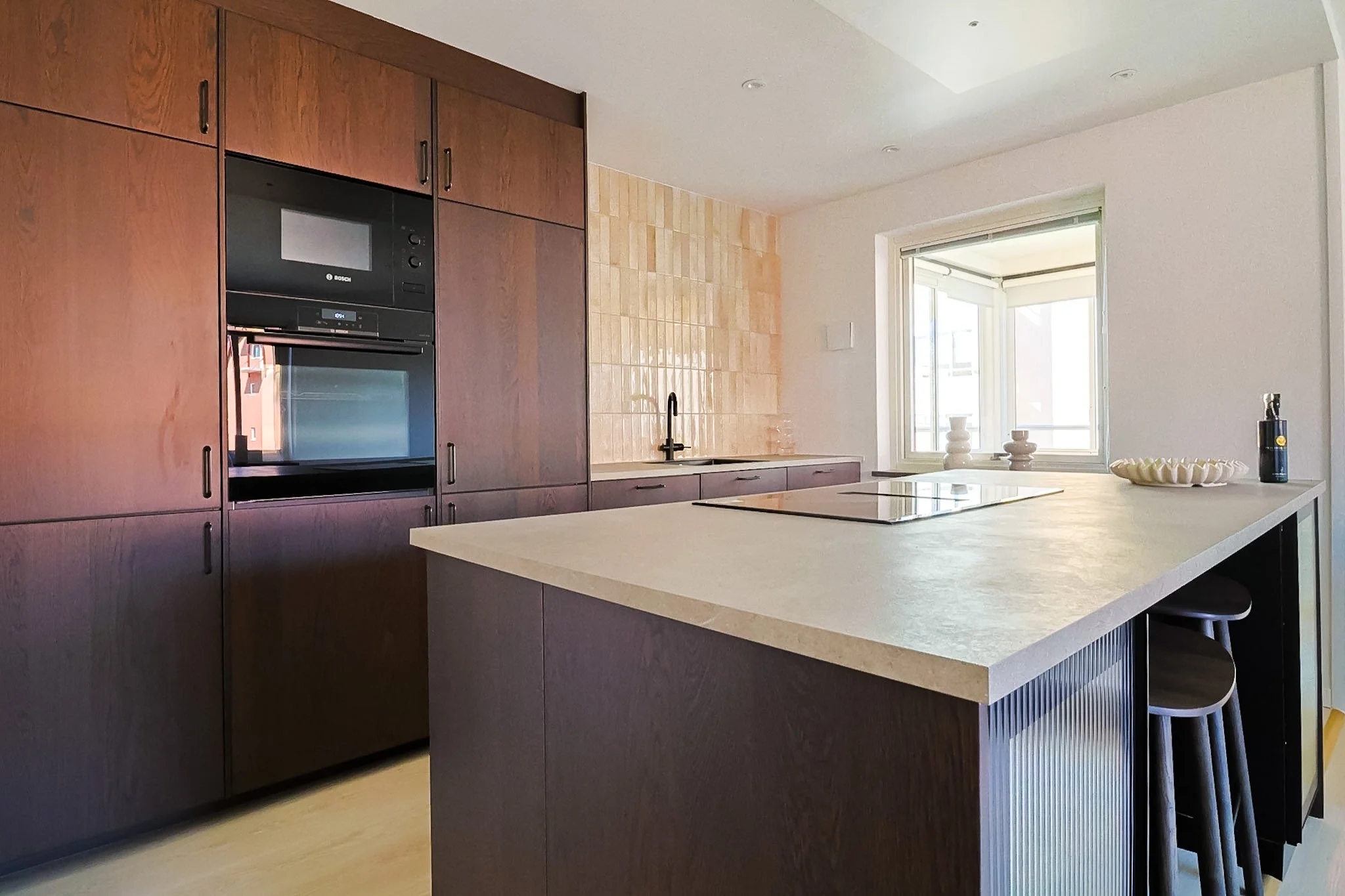 A modern kitchen with dark wood cabinets, a built-in microwave and oven, a beige tile backsplash, a white kitchen island with bar stools, and a large window with a view outside.