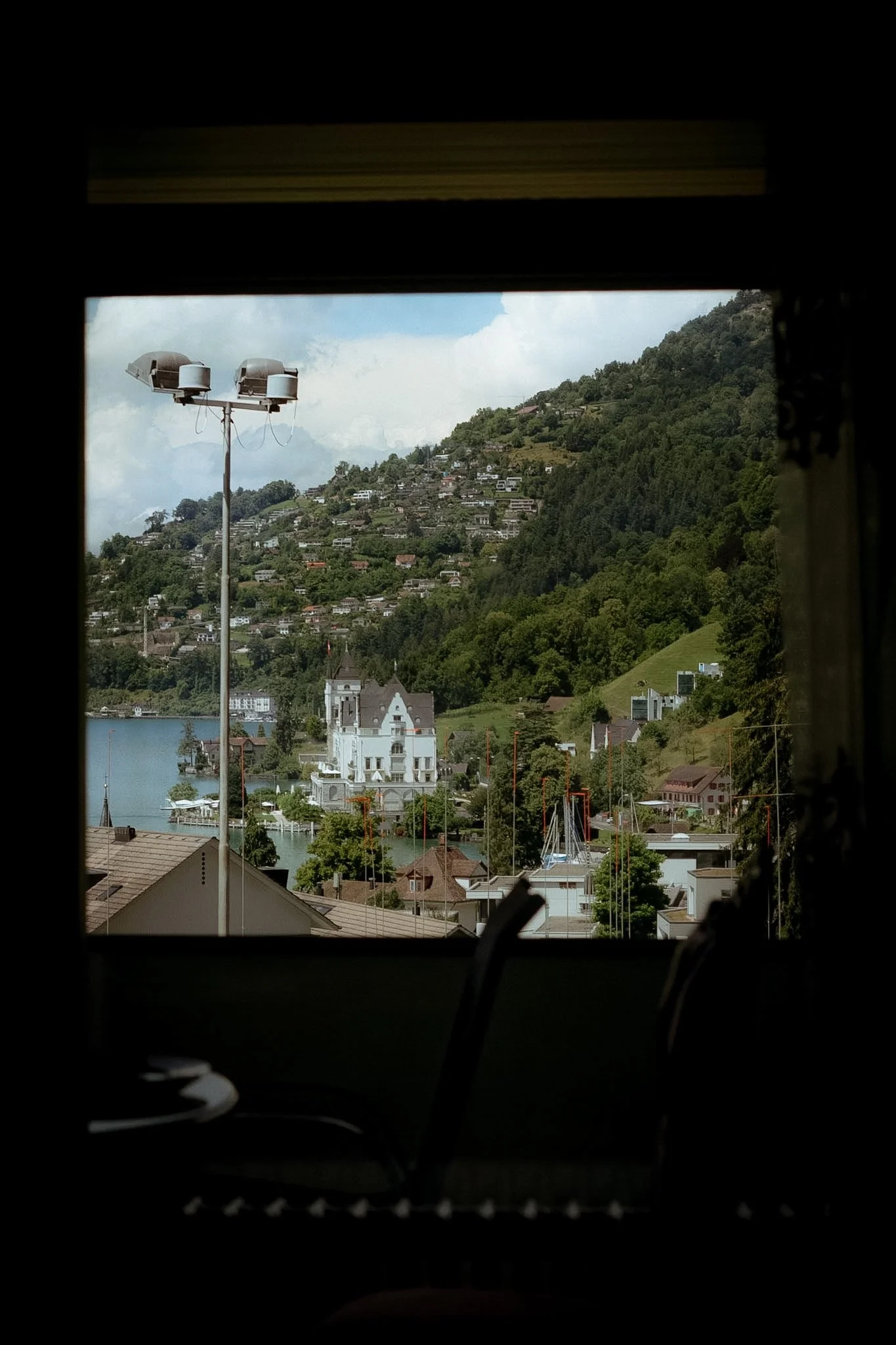 View of a hillside town with houses and trees, seen through a window with a dark frame, a streetlight with speakers, and a body of water in the foreground.