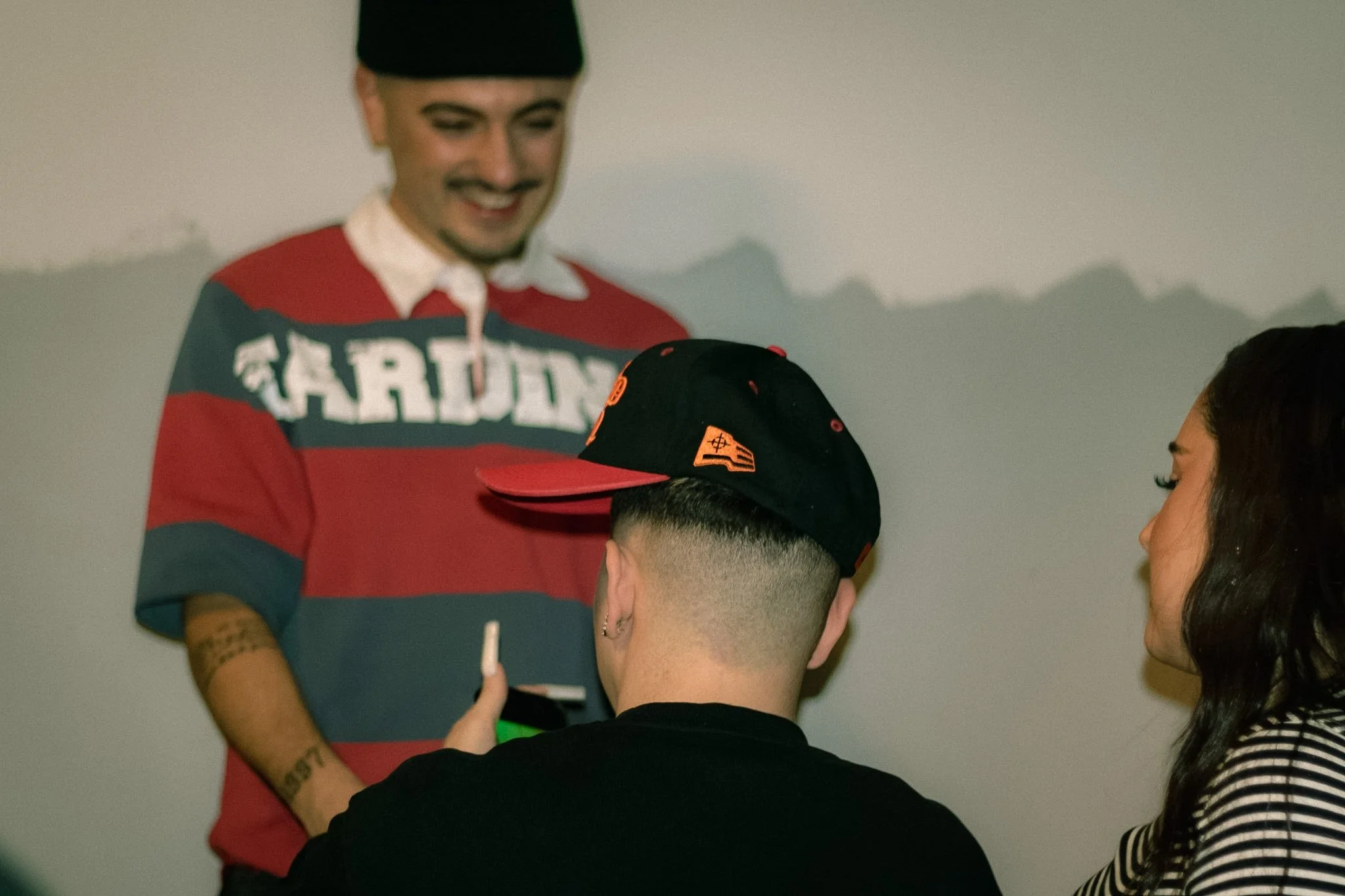 A group of people gathering, with one person smiling and wearing a squadron shirt, while others are engaged in conversation, indoors against a plain wall.