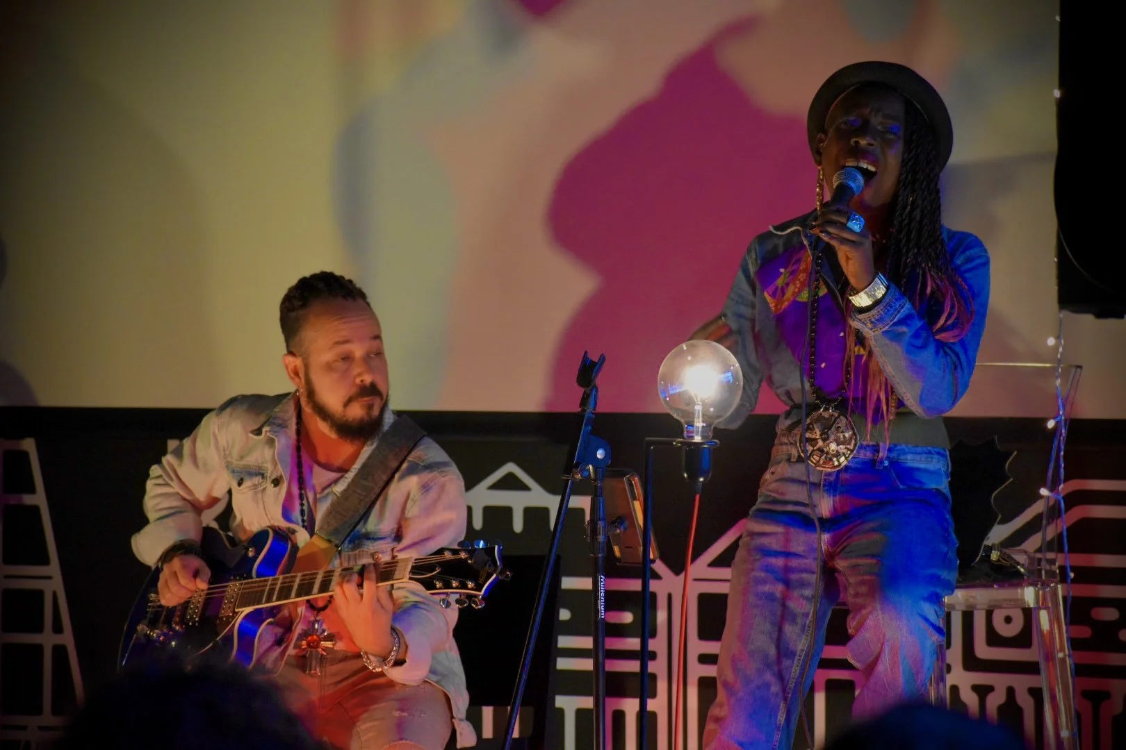 A woman singing into a microphone while a man plays guitar beside her. They are performing on a stage with colorful lighting and a large screen in the background.