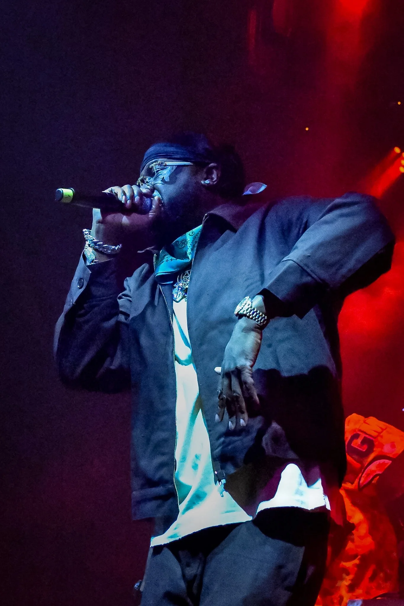 A performer holding a microphone on stage, wearing sunglasses, a bandana, gold jewelry, and casual clothing, with red stage lighting in the background.