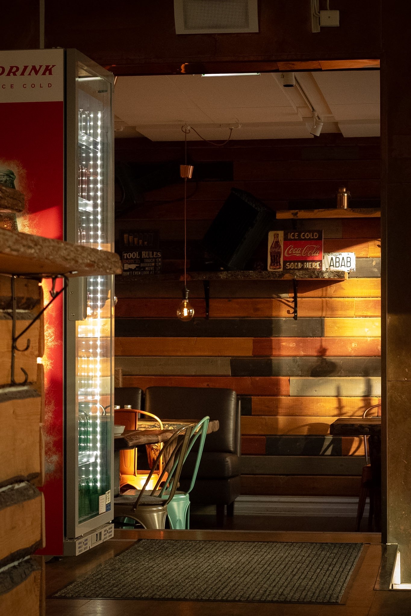 Interior of a cozy café or restaurant with wooden wall panels, a hanging light bulb, and various signs including a Coca-Cola advertisement and a license plate reading 'ABAB'.