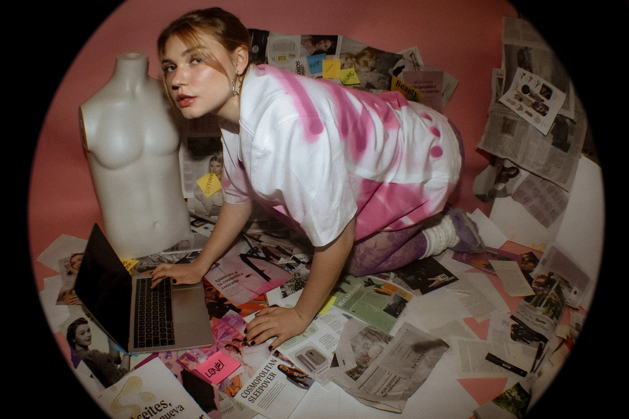 A woman with short hair and earrings is lying on a pink surface, surrounded by scattered magazines, papers, and clippings, with a mannequin torso nearby.