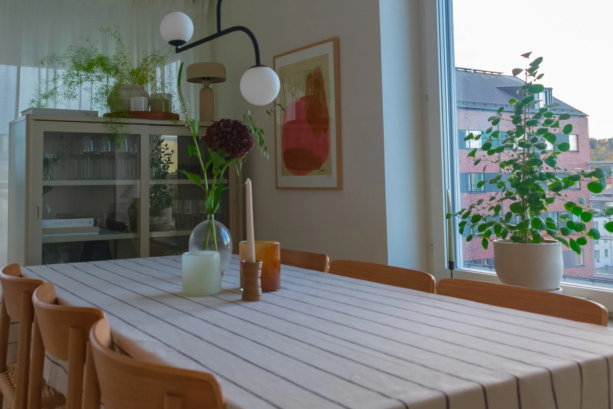 A dining room with a wooden table, chairs, potted plants on a cabinet and window sill, modern artwork on the wall, and large windows showing a brick building outside.