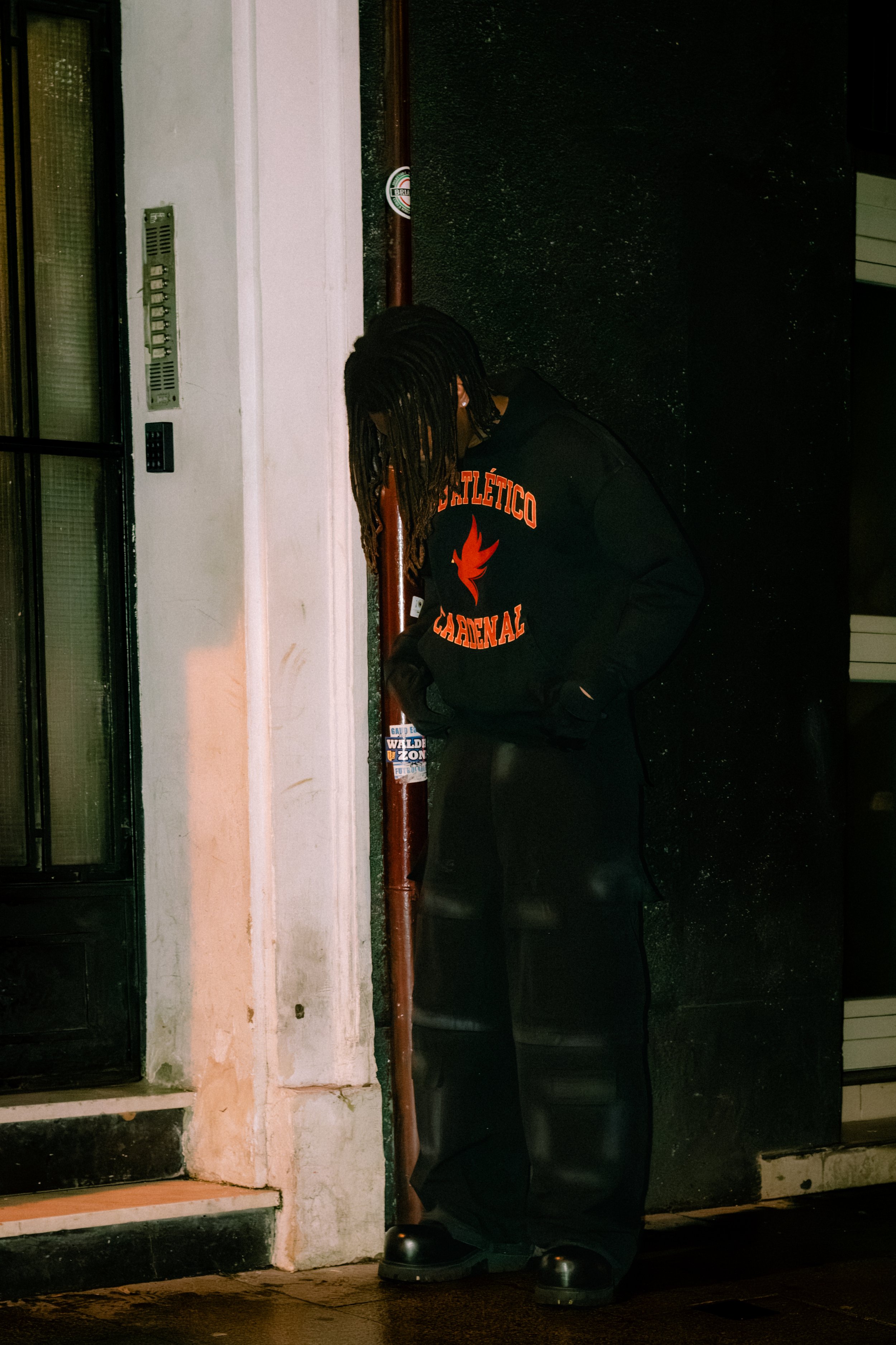 Person with long dreadlocks wearing a black hoodie with red and orange lettering, black pants, and large black shoes, standing outside against a black wall, looking downward, leaning against a painted white wall and a red pipe.