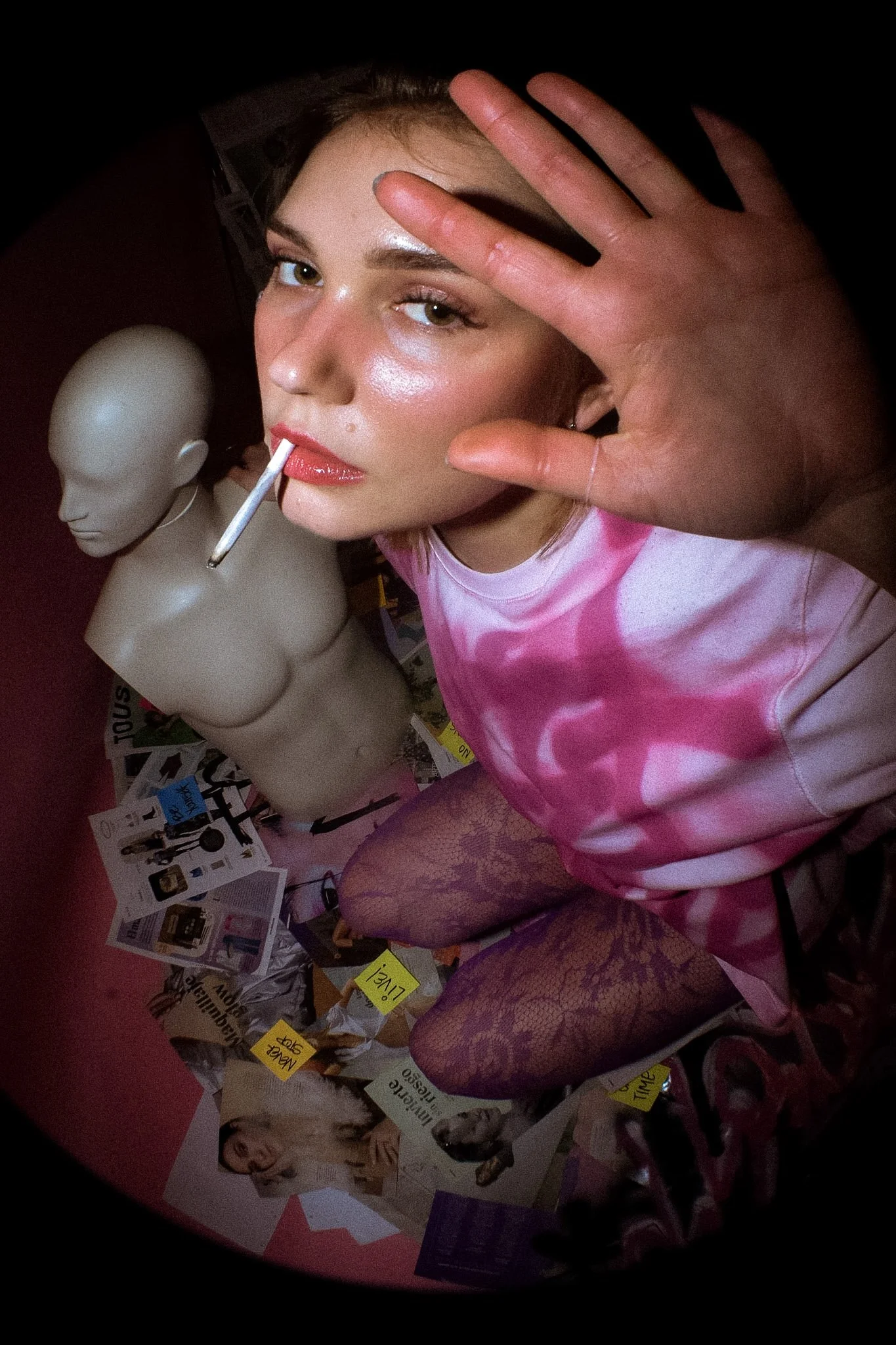 A person in pink tie-dye shirt and lace tights is near a mannequin head with a cigarette in its mouth, surrounded by magazines and notes on a wall.